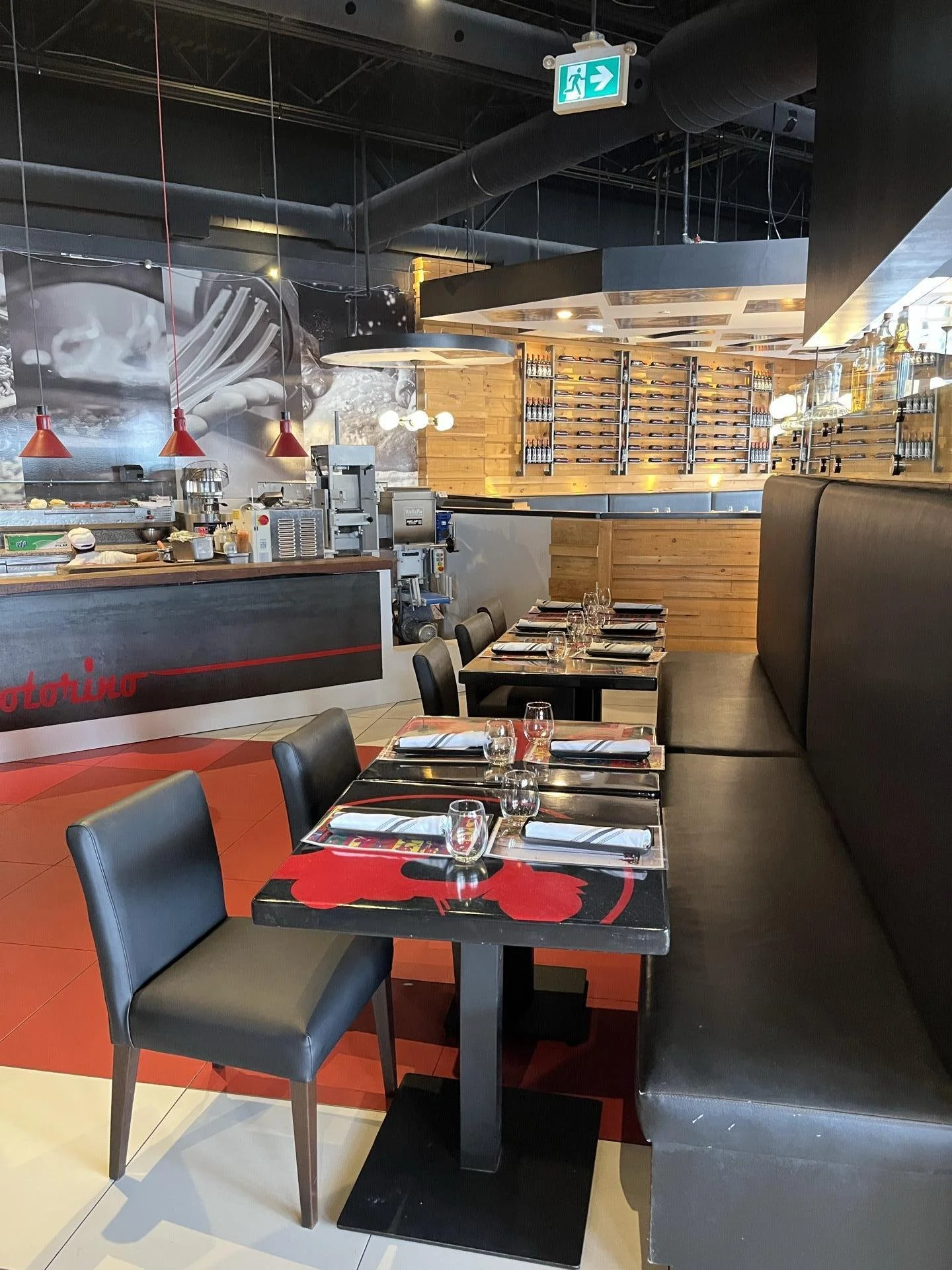 Interior of a modern restaurant with black tables, black chairs, and a long black booth. The tables are set with wine glasses, water glasses, and napkins. On the wall behind the tables, there is a wooden wall with wine bottles on display. The bar are
