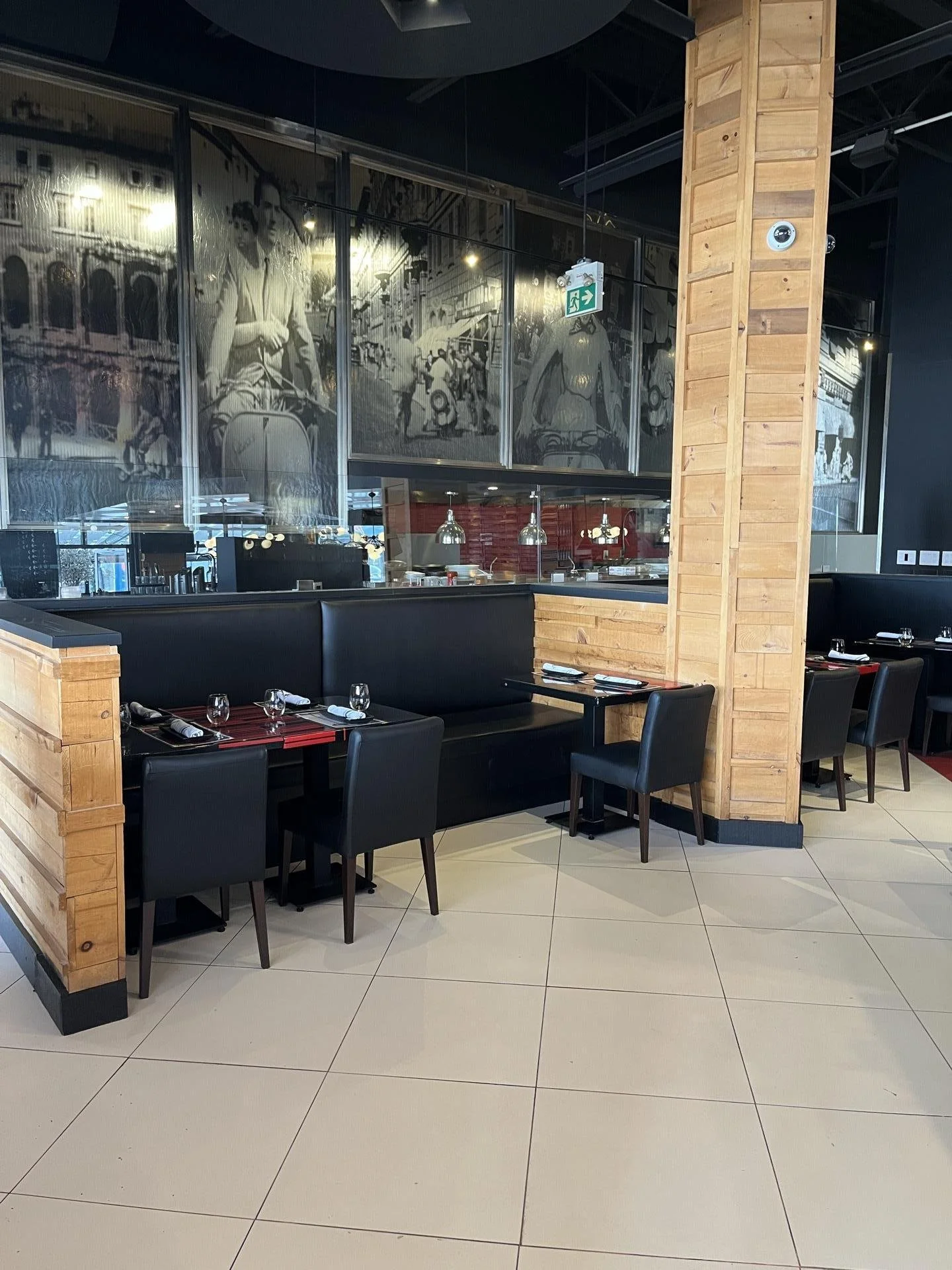 Inside a restaurant with black cushioned booths and black chairs, set tables with wine glasses and napkins, wooden paneled walls, and a large black-and-white mural of people on a street scene.