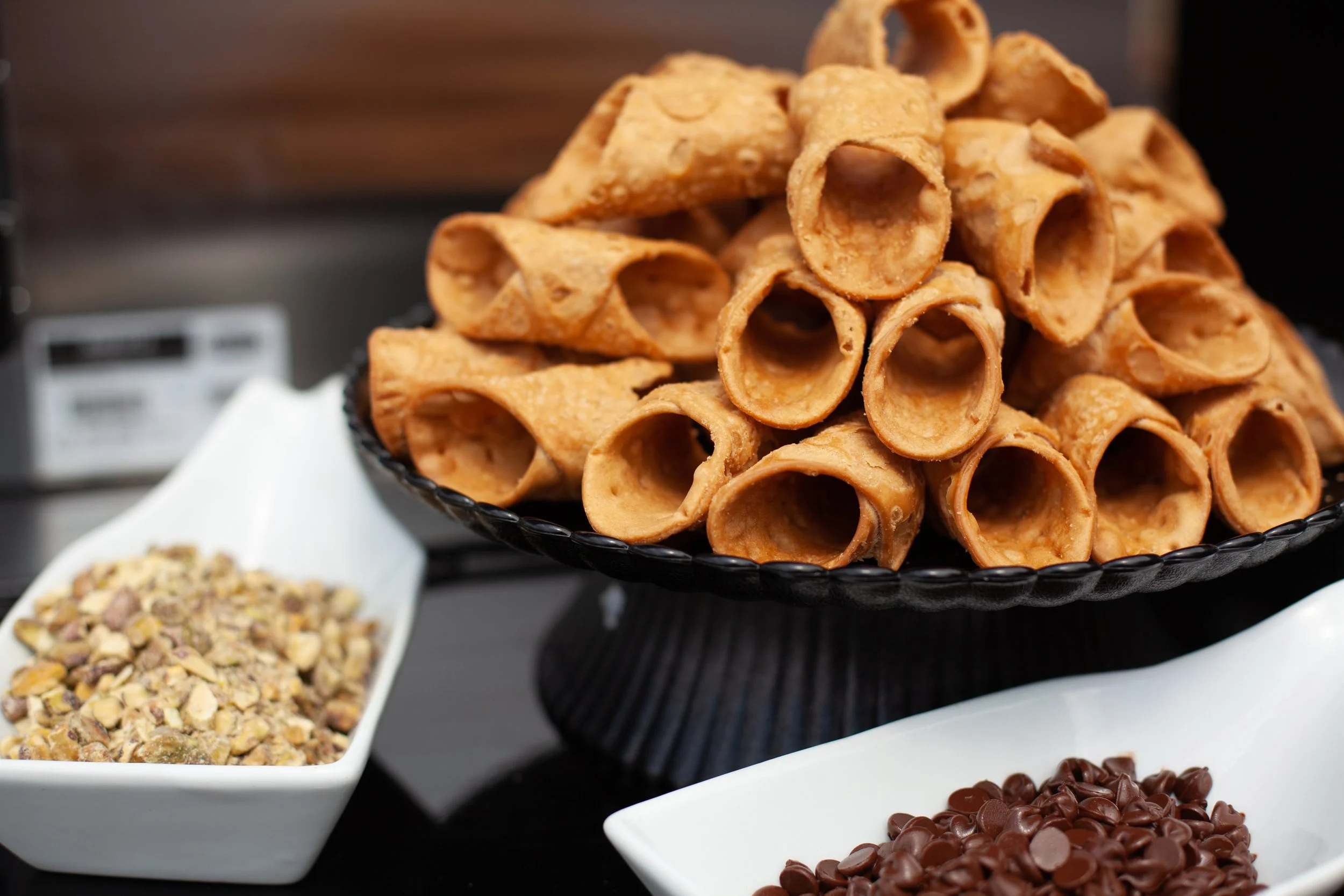 A black platter stacked with waffle cones, with bowls of chopped nuts and chocolate chips nearby.