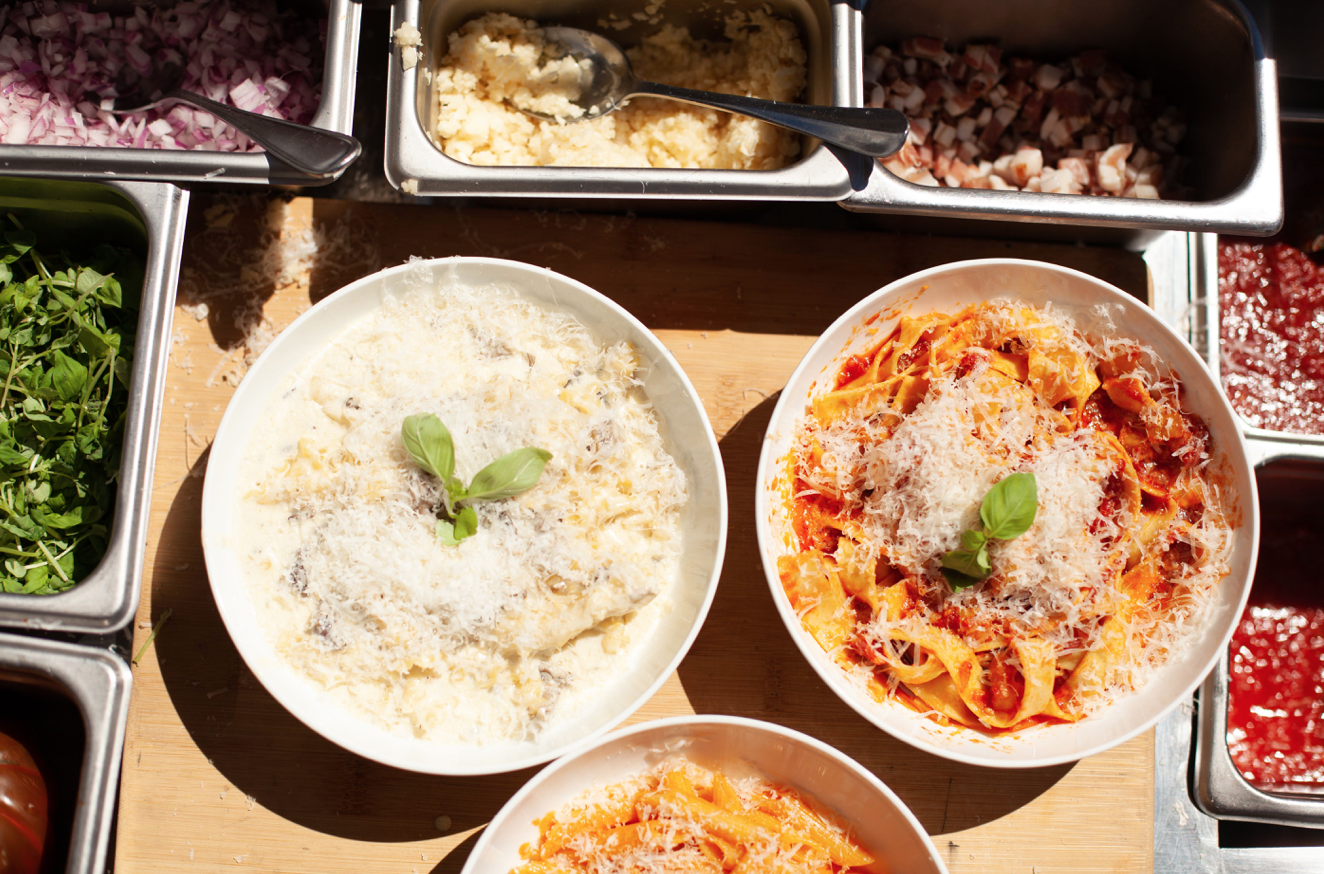Three plates of pasta with cheese and basil garnishes, surrounded by containers of chopped vegetables, garlic, diced bacon, and pasta sauces.