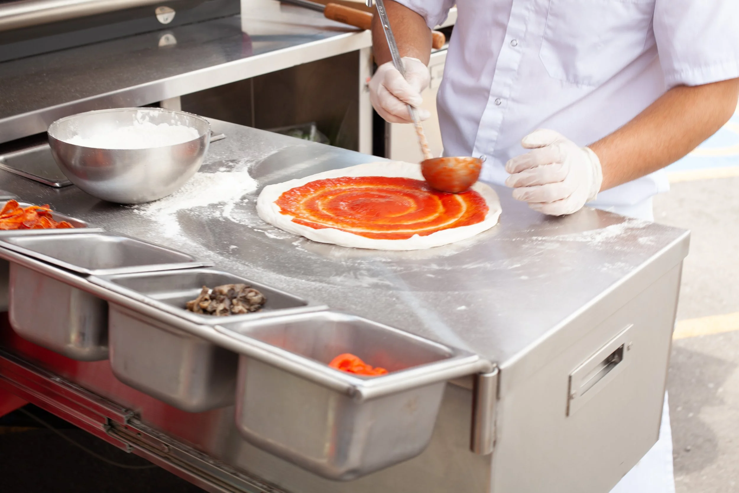 A person in a white shirt and gloves preparing a pizza on a stainless steel counter, spreading tomato sauce on dough with a ladle, with various ingredients in small containers nearby.