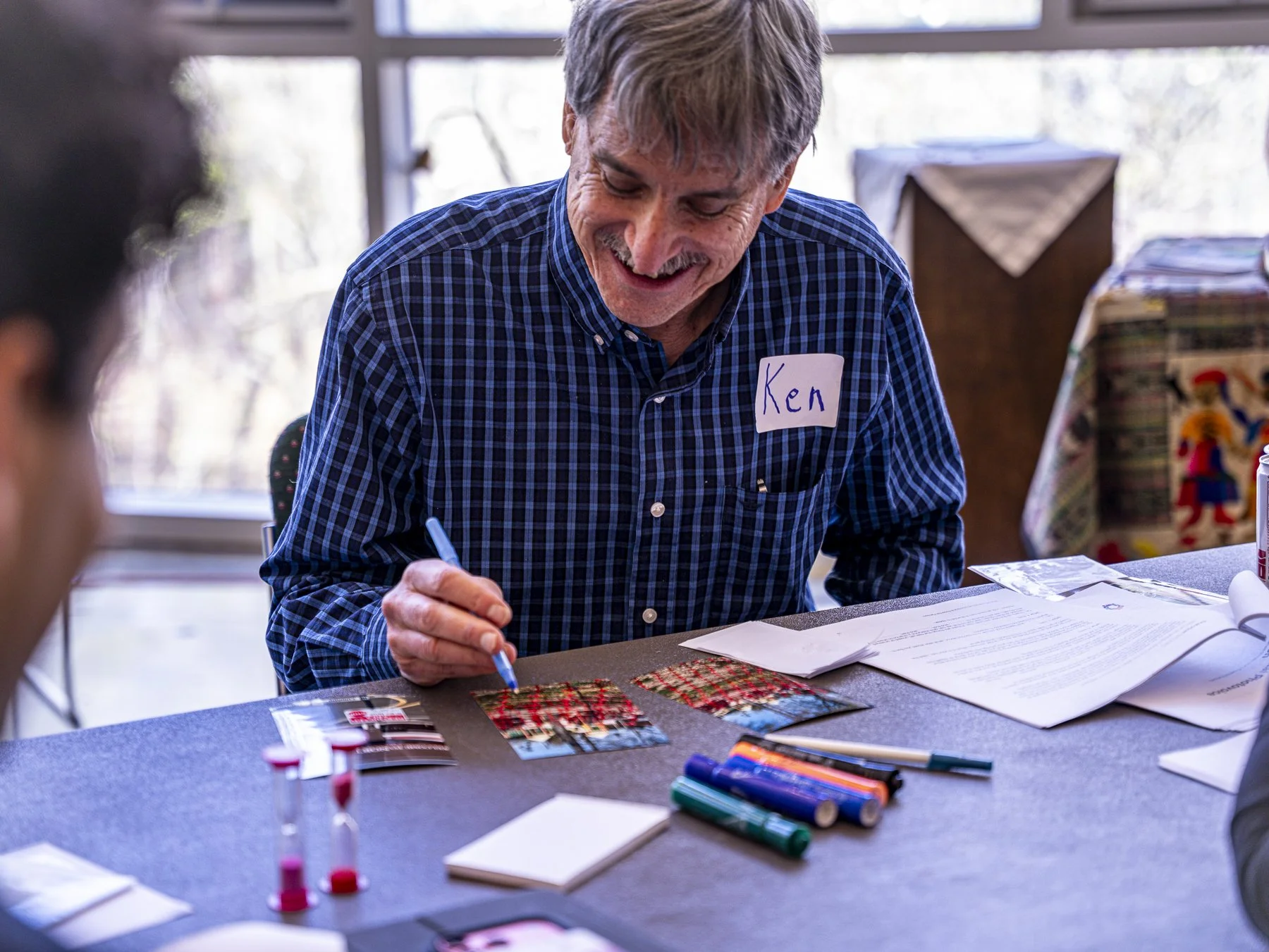 A smiling man with gray hair, wearing a blue plaid shirt with a name tag labeled 'Ken,' sitting at a table covered with documents, copy paper, markers, and holiday-themed fabric samples, in a bright room with large windows.
