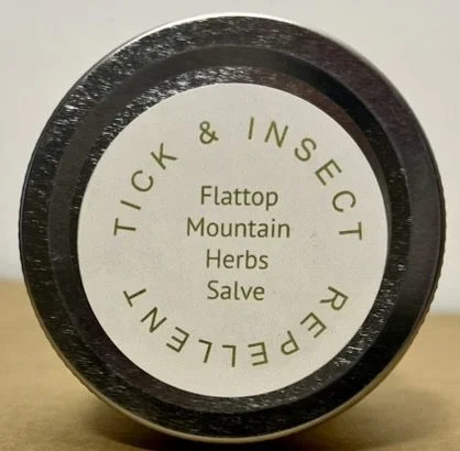 Close-up of a black container lid with a cream label that reads: 'Tick & Insect Repellent Flattop Mountain Herbs Salve'.