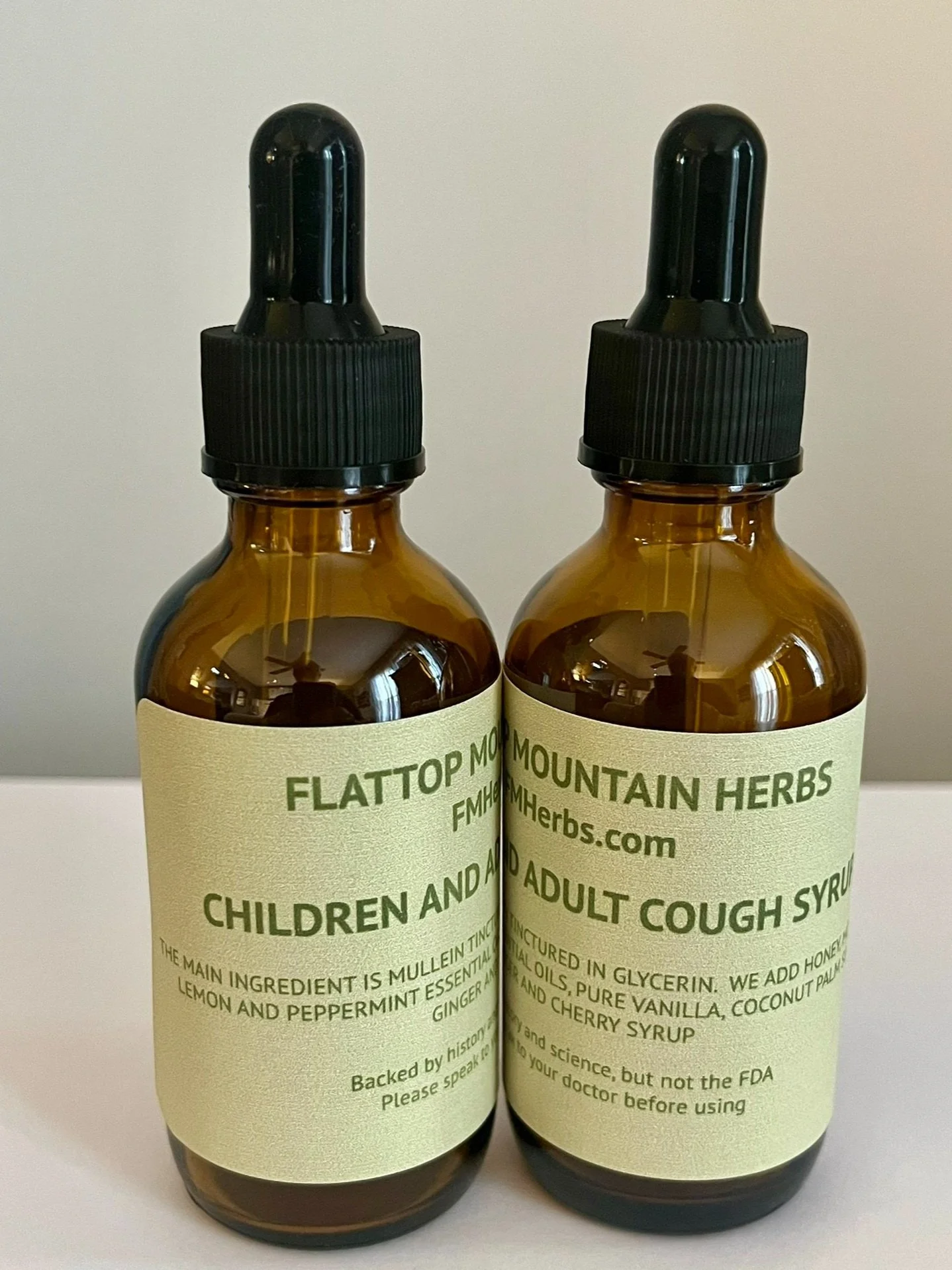 Two amber glass dropper bottles with black caps and labels reading 'Flattop Mountain Herbs' containing herbal cough syrup for children and adults.