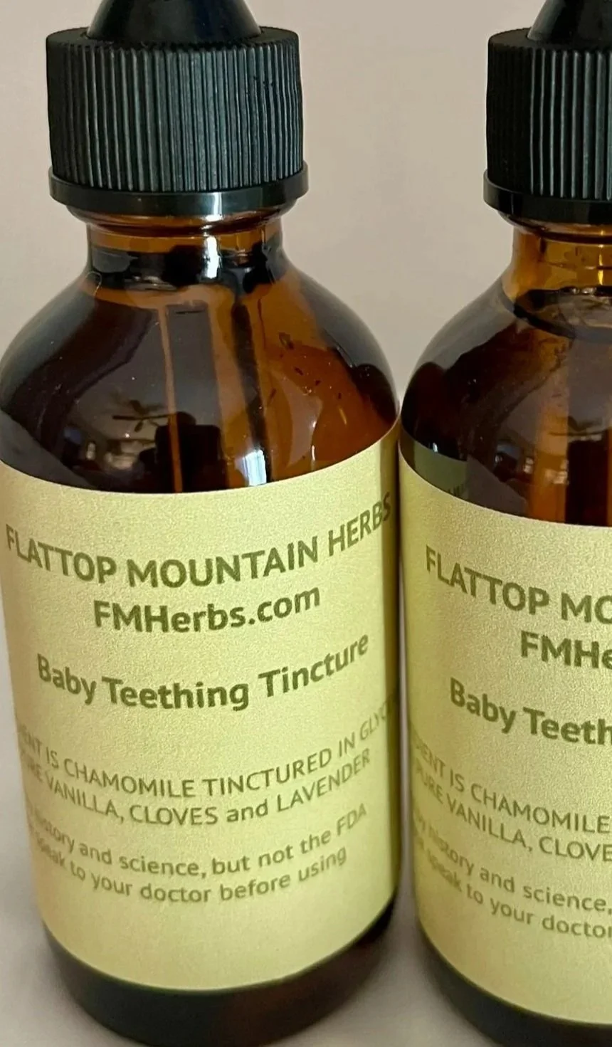 Two brown glass bottles with black caps, labeled as 'Baby Teething Tincture' from Flattop Mountain Herbs, with a cream-colored label indicating ingredients including chamomile, vanilla, cloves, and lavender.