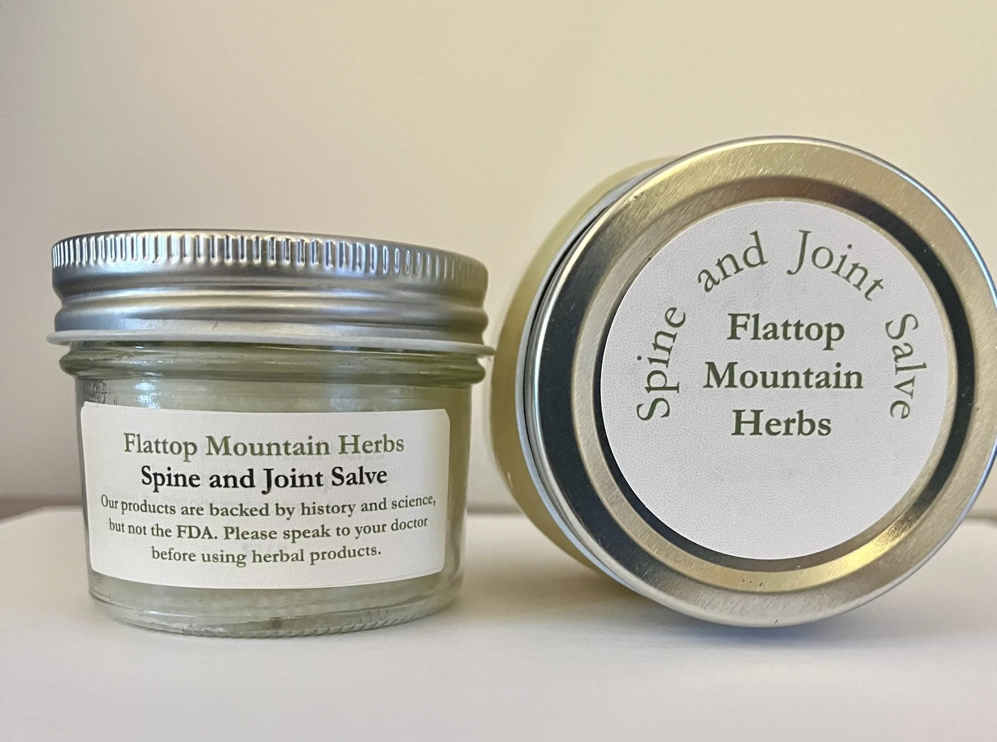 Close-up of herbal products, including a small jar with a white label and a silver lid, and a round metal tin with a white label. Both labels mention 'Flatop Mountain Herbs' and 'Spine and Joint Salve'. The tin lid is partially visible with a gold-co