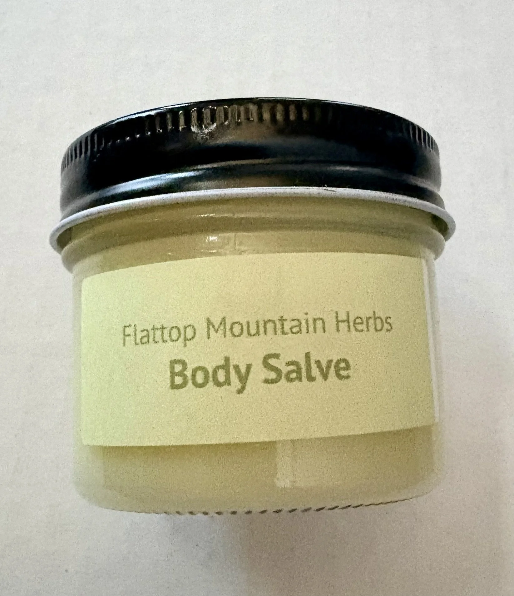 Close-up of a container of body salve labeled 'Flatrop Mountain Herbs Body Salve' with a black lid, on a white surface.
