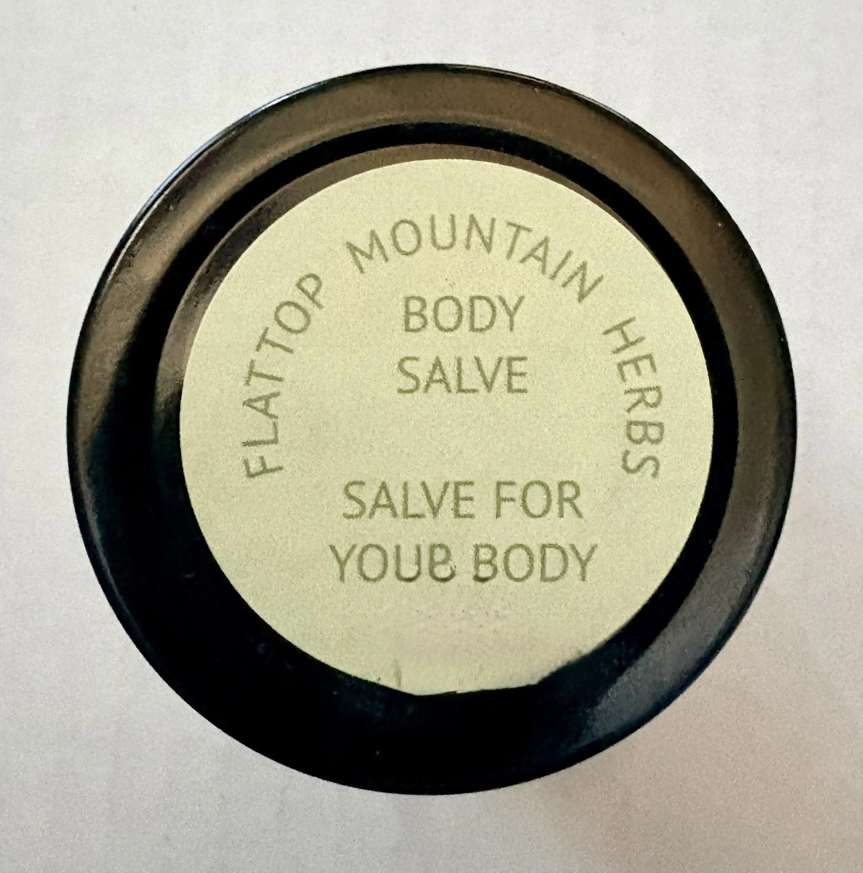 A round container with a label that reads 'FACIAL TOP MOUNTAIN HERBAL BODY SALVE' and additional text about salve for your body.