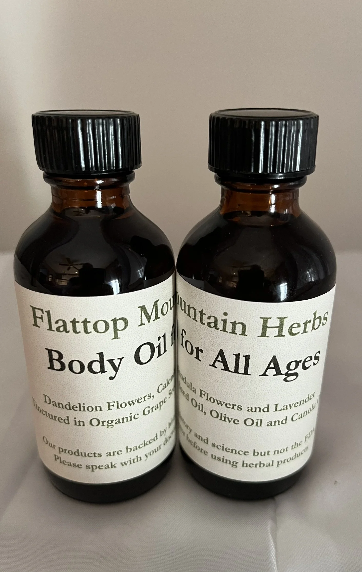 Two dark amber bottles of body oil with white labels and black caps, placed side by side against a plain background.