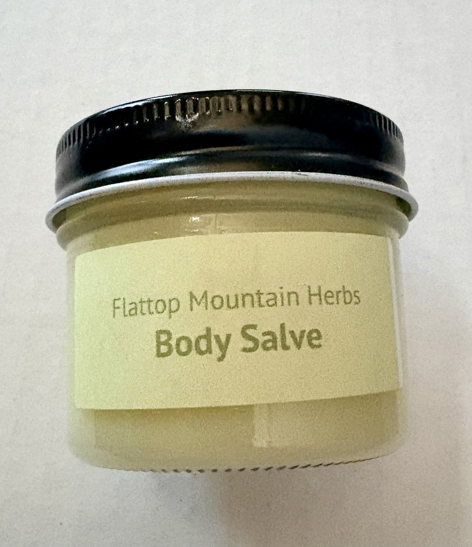 Close-up of a container with a black lid labeled 'Flatop Mountain Herbs' and 'Body Salve' in yellowish-green packaging.
