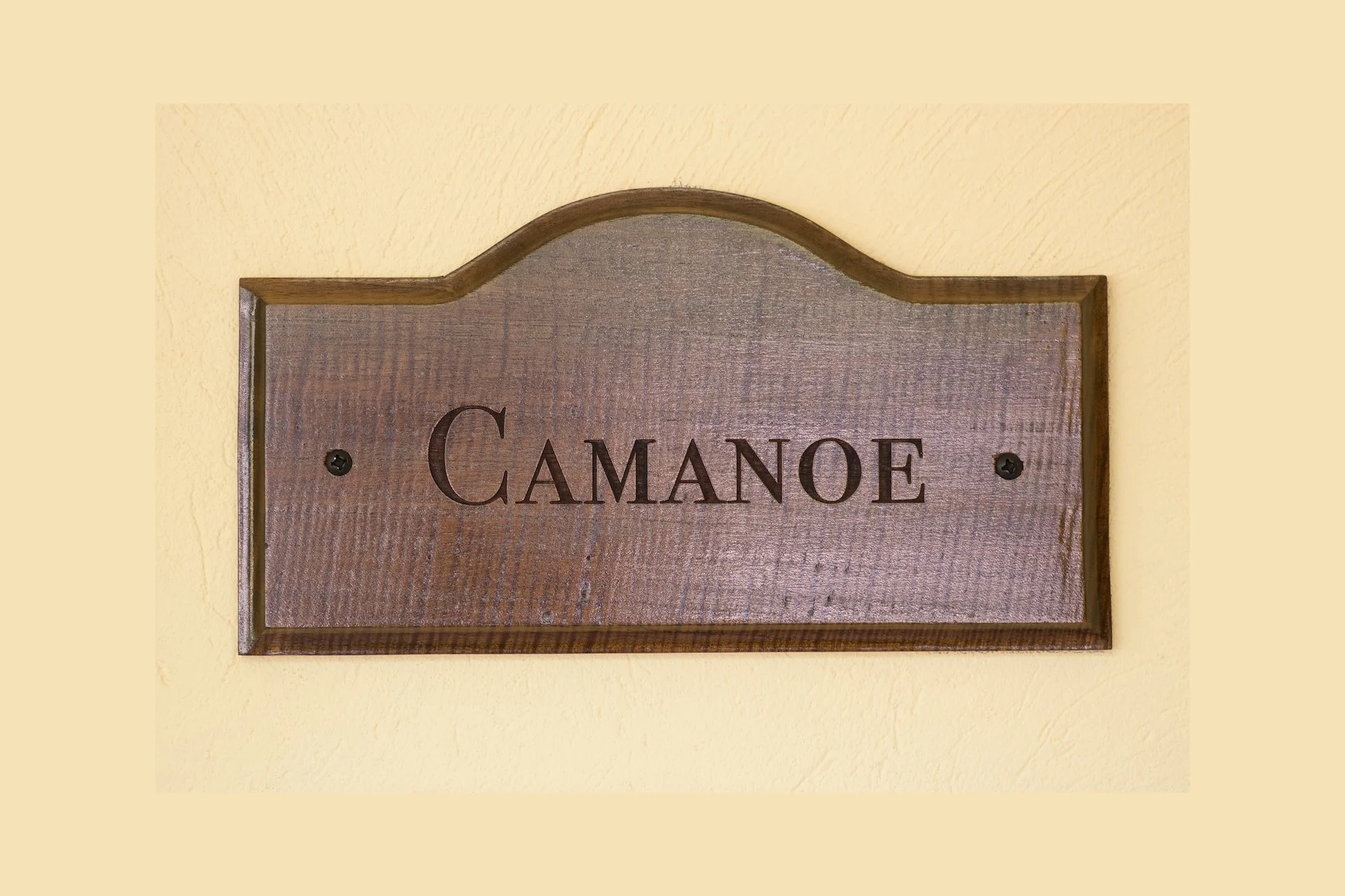 Wooden sign with the word 'CAMANOE' engraved, mounted on a yellow wall.