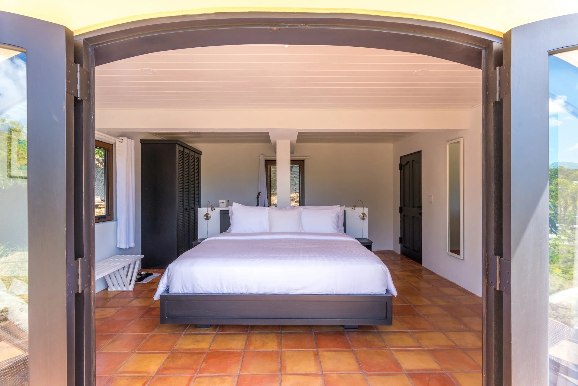 Modern bedroom with white bedding, dark wood wardrobe, and terracotta floor tiles viewed through open double doors.