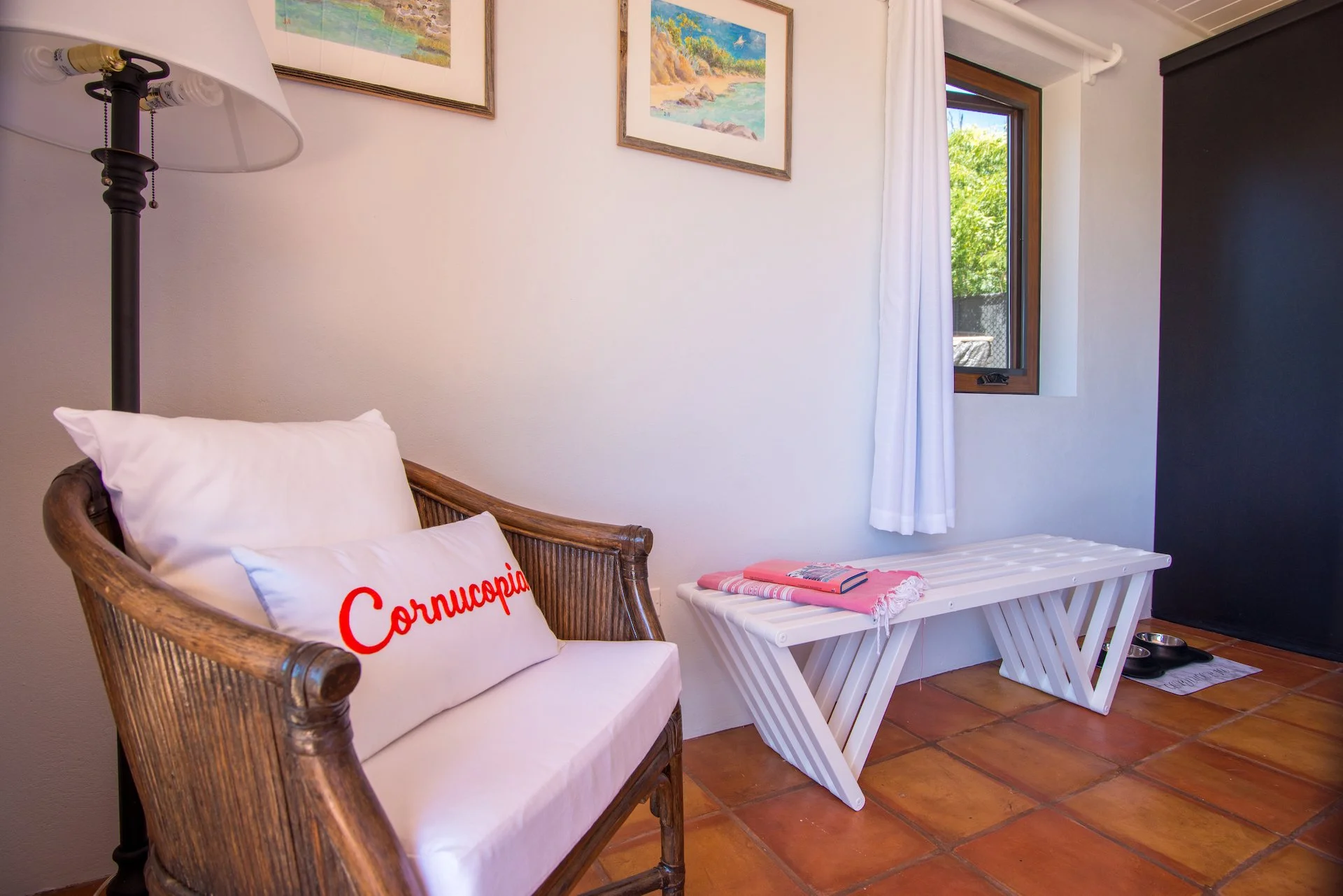 Cozy interior with wicker chair, Cornucopia pillow, floor lamp, white bench under window, towels, and books on terracotta tile floor.