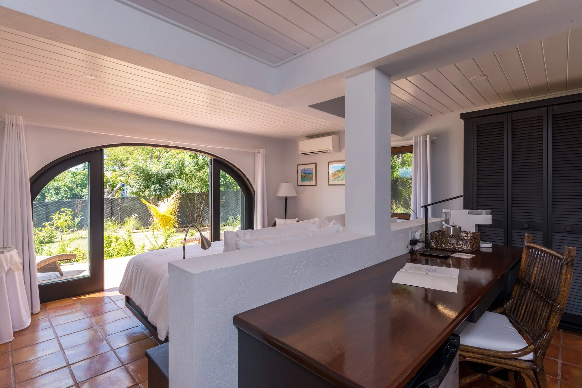 Modern bedroom with large arched windows, white walls, wooden desk, and patio view.