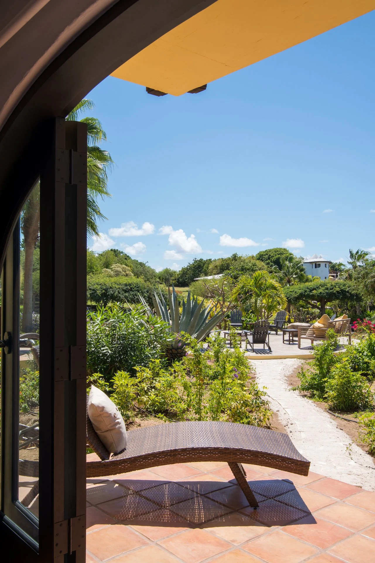 Outdoor patio view with a wicker lounge chair, terracotta tile flooring, tropical plants, and garden seating under a clear blue sky.