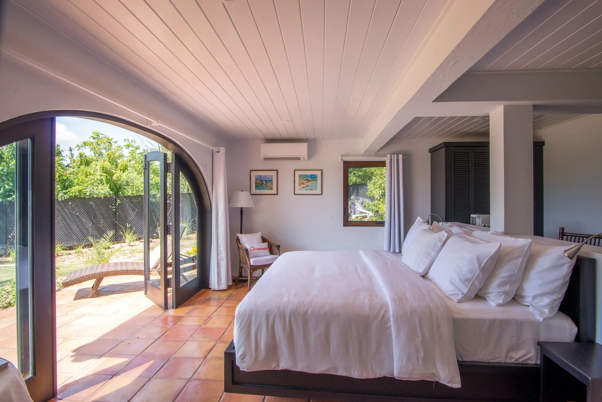 Bright bedroom with white bedding, large arched glass doors opening to a patio, terracotta tile floor, wicker chair with a cushion, table lamp, and wall art. Outdoor greenery visible through windows.