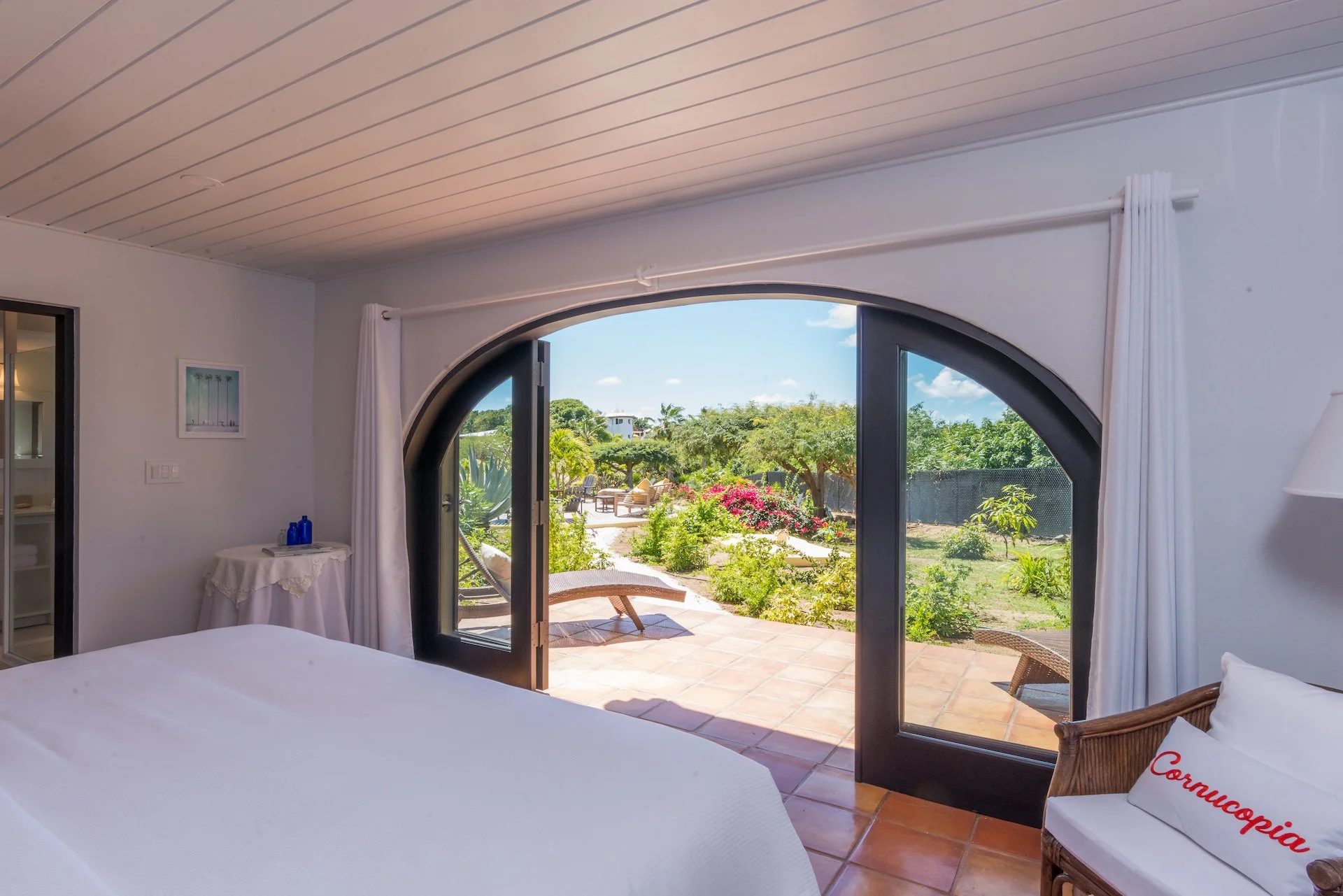 Bedroom interior with arched glass doors opening to a garden, featuring a bed, white curtains, and outdoor lounge chairs.
