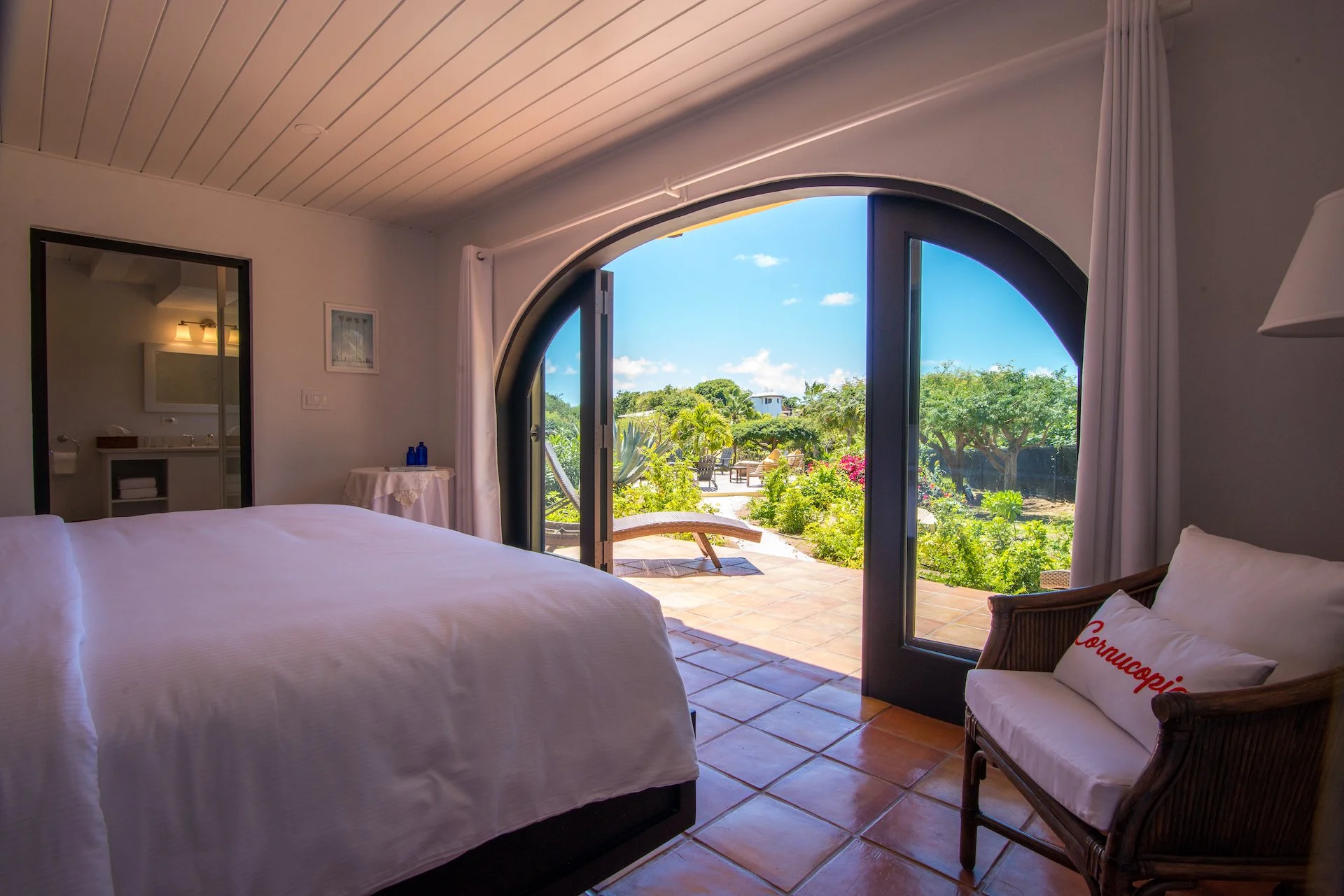 Bedroom with white bed, open glass doors leading to a sunny outdoor patio with garden view, wicker chair, and adjacent bathroom visible.