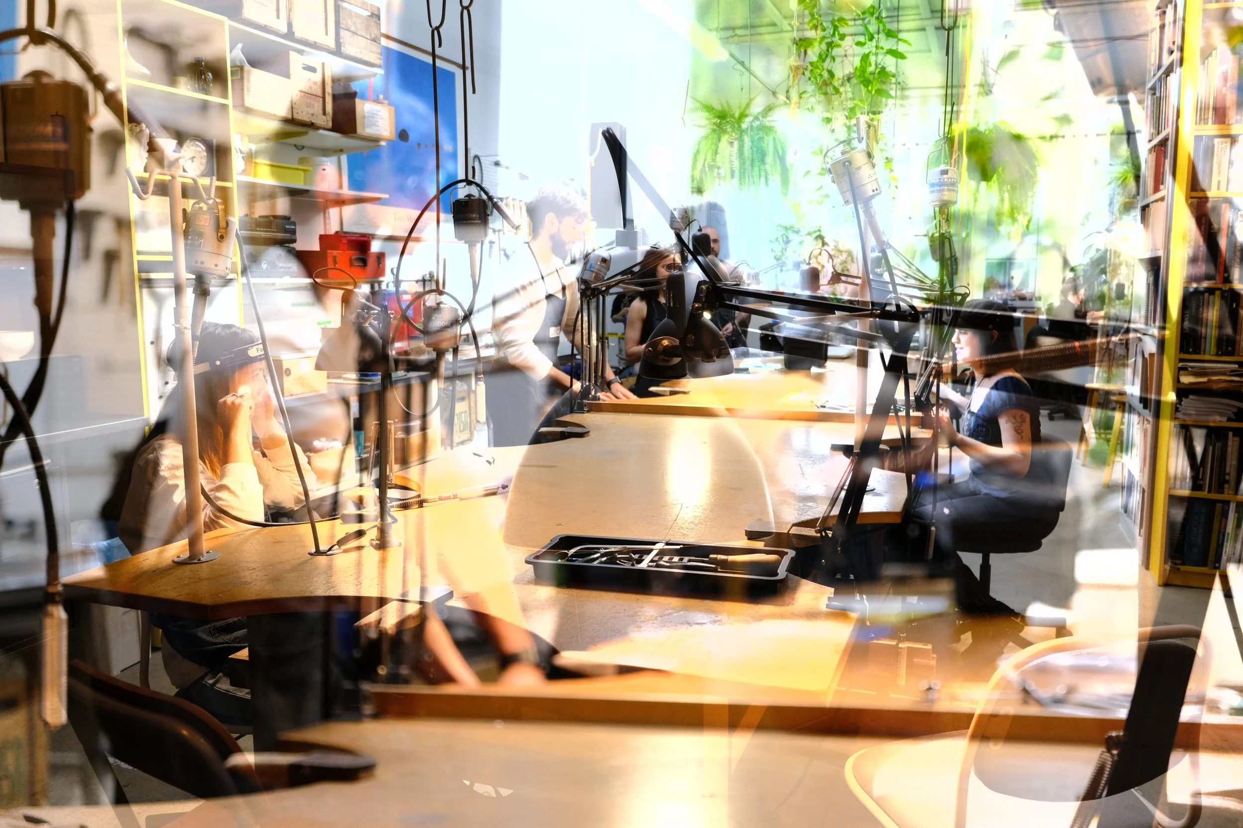 A group of people working in a studio with a large wooden table, surrounded by bookshelves, plants, and various studio equipment, visible through a glass window.