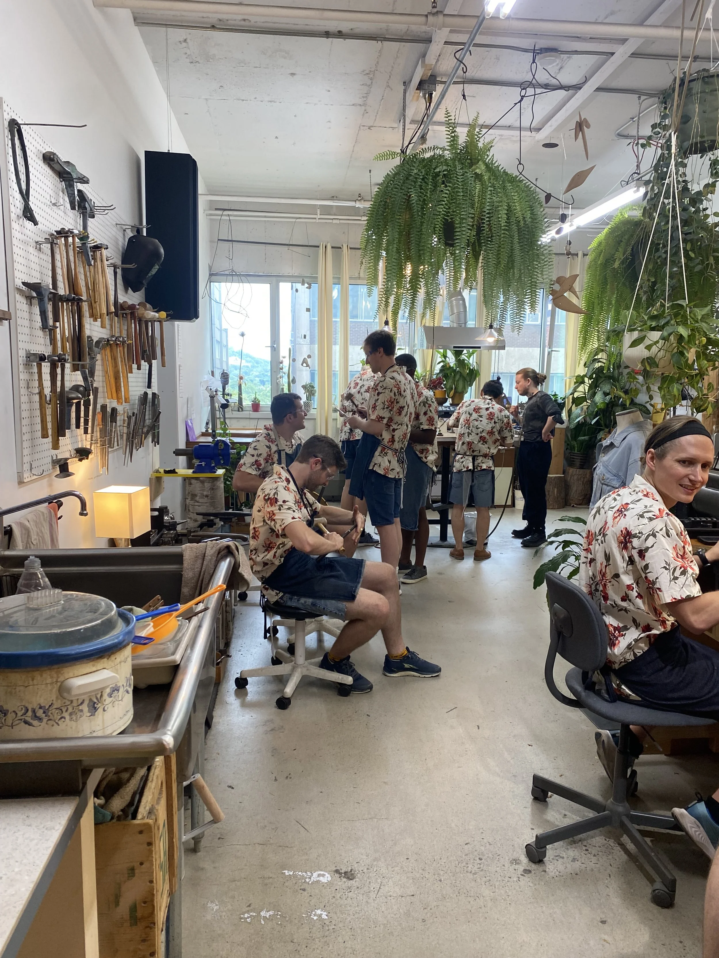 Des personnes dans un atelier ou une salle de classe portant des chemises hawaïennes similaires, certaines travaillant à des tables et d'autres debout, avec des outils accrochés au mur et des plantes suspendues au plafond.
