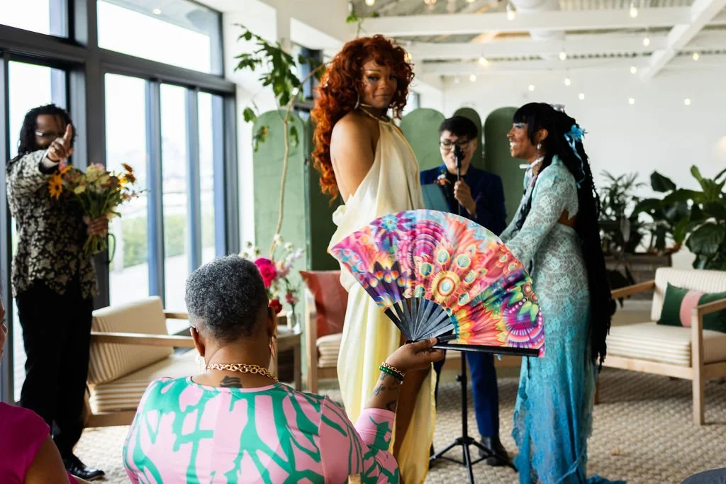 A queer, trans couple during their wedding ceremony presided by queer officiant from Once Upon A Vow in Brooklyn Grange. Parter in yellow gown is looking at mom in front row with colorful fan out while the other partner is smiling at them.
