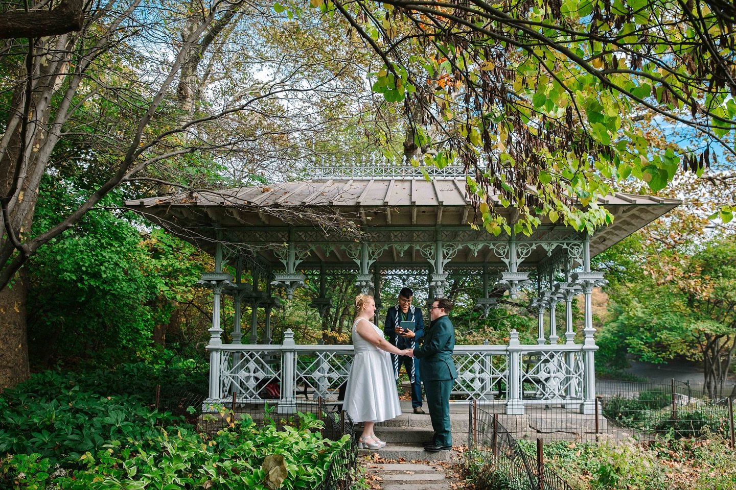 Trans couple, partner in a forest green suit holds hands with partner in white wedding dress during their Central Park elopement ceremony at Ladies Pavilion with queer wedding officiant from Once Upon A Vow in the middle