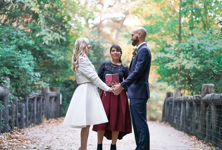 A couple holding hands during their outdoor NYC elopement, bride in white dress and groom in black tux, with their Brooklyn wedding officiant in between from Once Upon A Vow marrying them in Prospect Park's Binnen Bridge