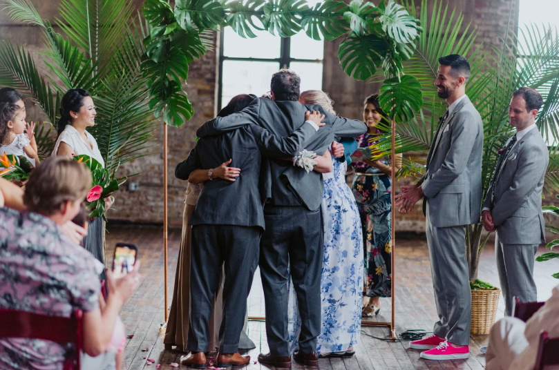 Beneath a montesera wedding arch, there are two men in gray suits and two women beside them all embracing during a ring warming ceremony held at Greenpoint Loft officiated by Fun, Personal, Brooklyn Wedding Officiant from Once Upon A Vow