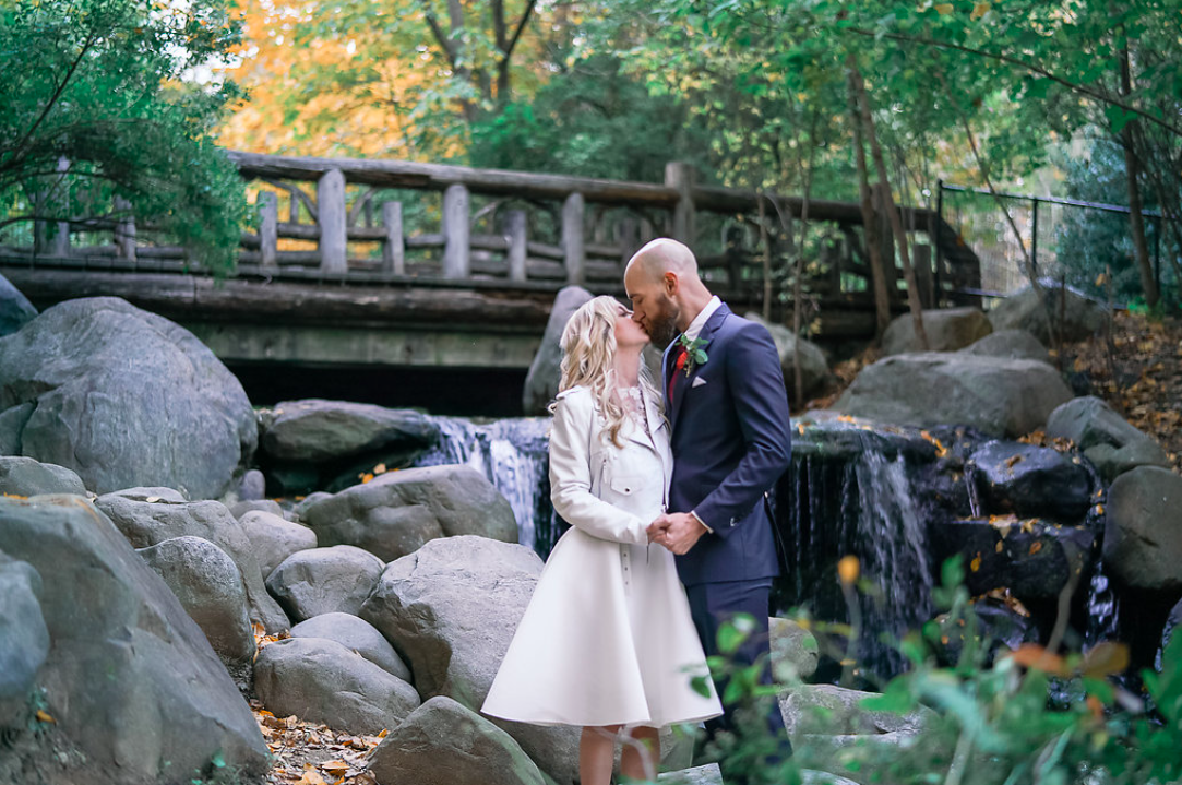 A couple kissing and holding hands, bride in white dress and groom in black suit, after their outdoor Brooklyn elopement officiated by Brooklyn wedding officiant from Once Upon A Vow in front of Prospect Park's Binnen Falls