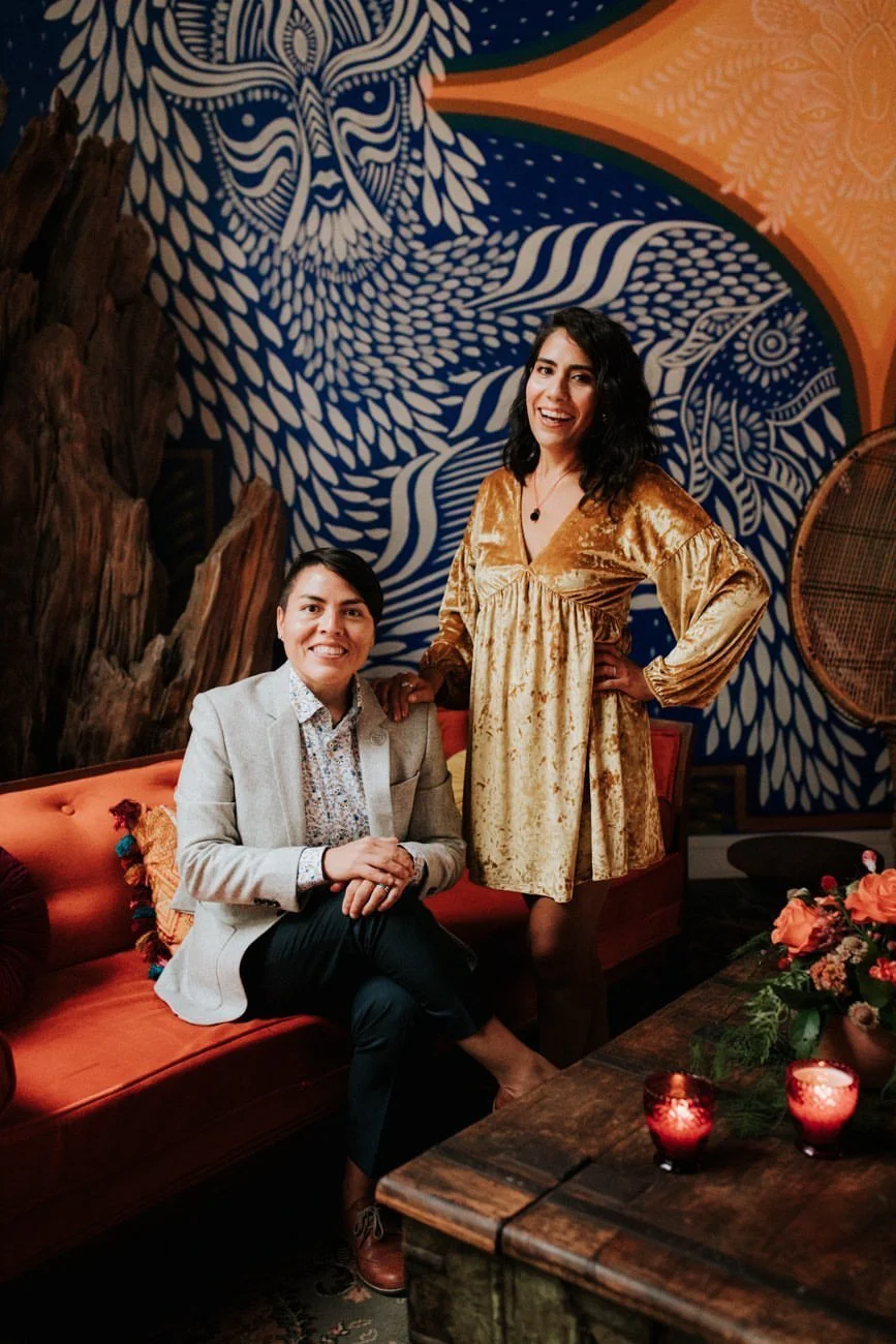 Formal photo of sibling bilingual wedding officiants from Once Upon A Vow, Karla sitting on orange couch wearing black pants, floral shirt, and light grey blazer with Dani standing wearing a yellow, velvet mini dress; vibrant blue mural background