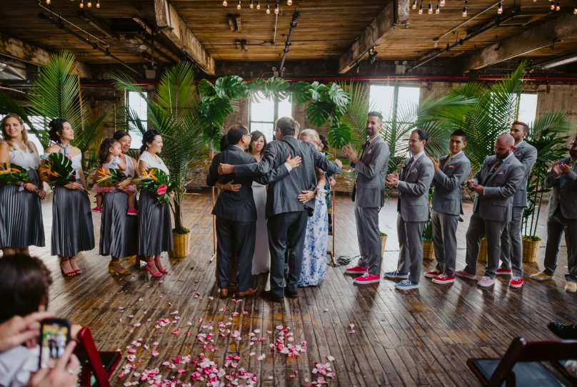 Beneath a montesera wedding arch, there are two men in gray suits and two women beside them all embracing during a ring warming ceremony held at Greenpoint Loft officiated by Fun, Personal, Brooklyn Wedding Officiant from Once Upon A Vow
