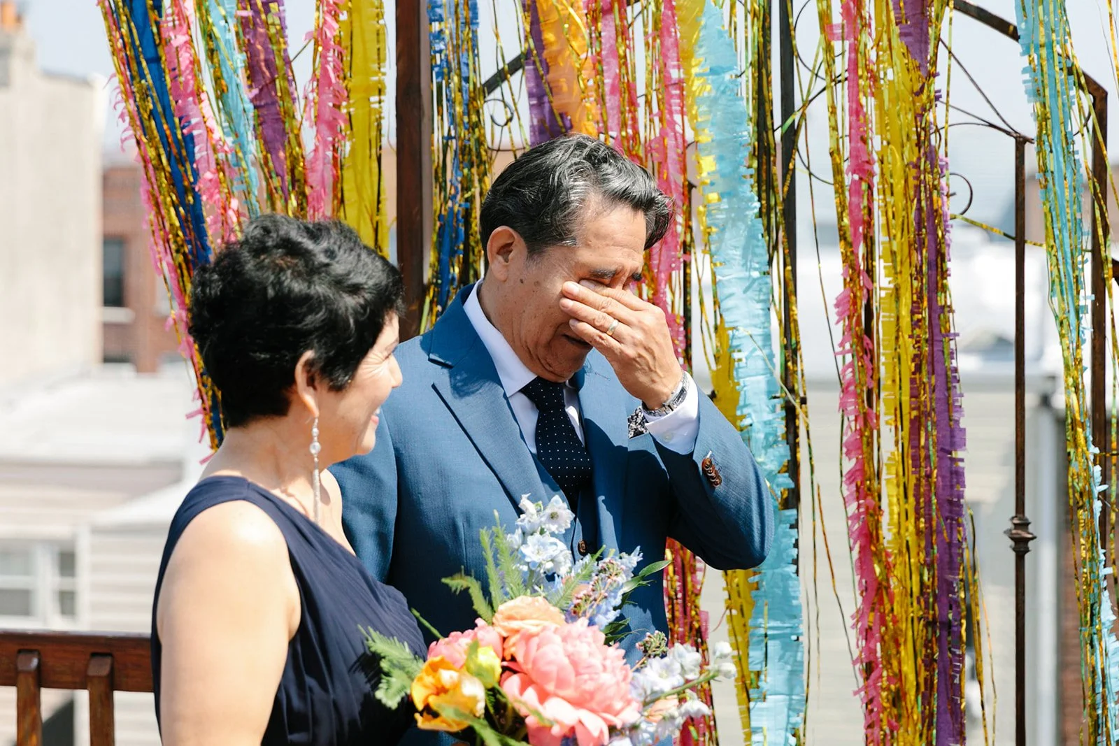 Older Latino man wiping away tears as wife with short hair stands beside him holding bouquet during their wedding ceremony vow renewal officiated by bilingual Brooklyn wedding officiant from Once Upon A Vow with colorful tassle backdrop