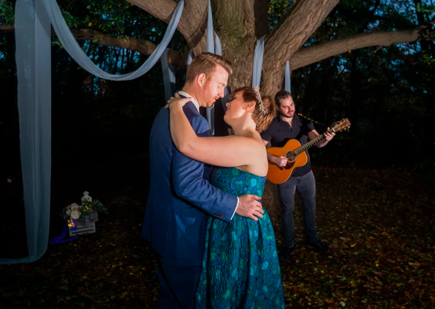 Man in blue suit and woman in green/blue dress dancing beneath beautiful large tree with guitarist in background after they said their wedding vows with fun Brooklyn Wedding Officiant from Once Upon A Vow