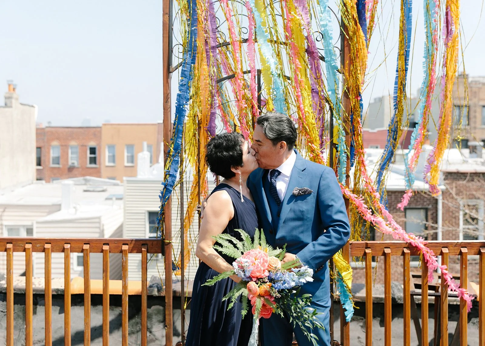 Couple in their 60's kissing in front of a colorful paper frill backdrop during their vow renewal officiated by bilingual wedding officiants from Once Upon A Vow, wife wears blue dress and holds colorful bouquet and husband wears a blue suiit