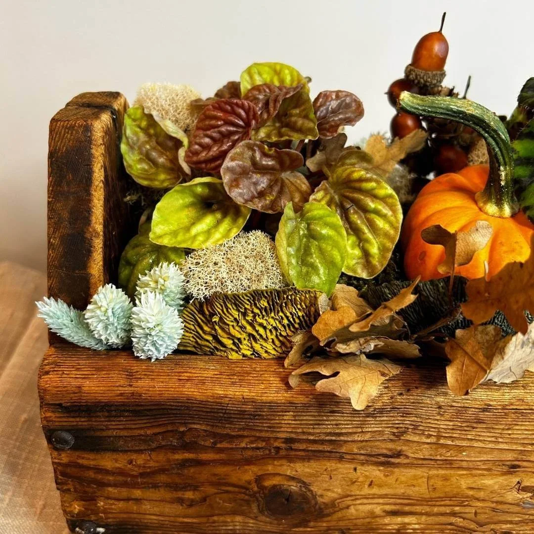 closeup of part of a thanksgiving table centerpiece with pumpkins peperomia leaves dried flowers mushrooms acorns on tan tablecloth