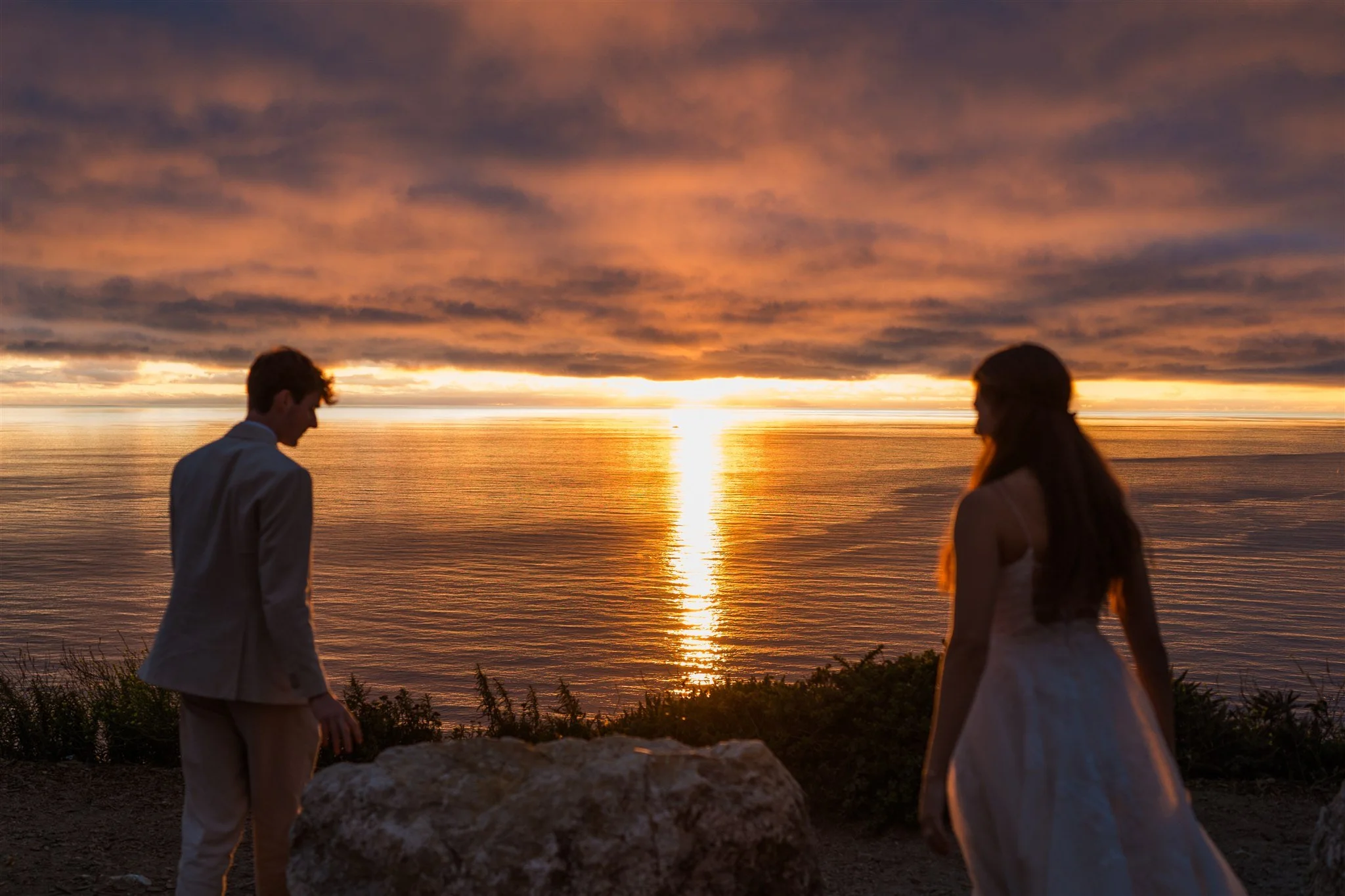 A couple dressed in formal attire stands near a large rock on a hillside overlooking the ocean during a vibrant sunset with colorful clouds and a glowing reflection on the water.