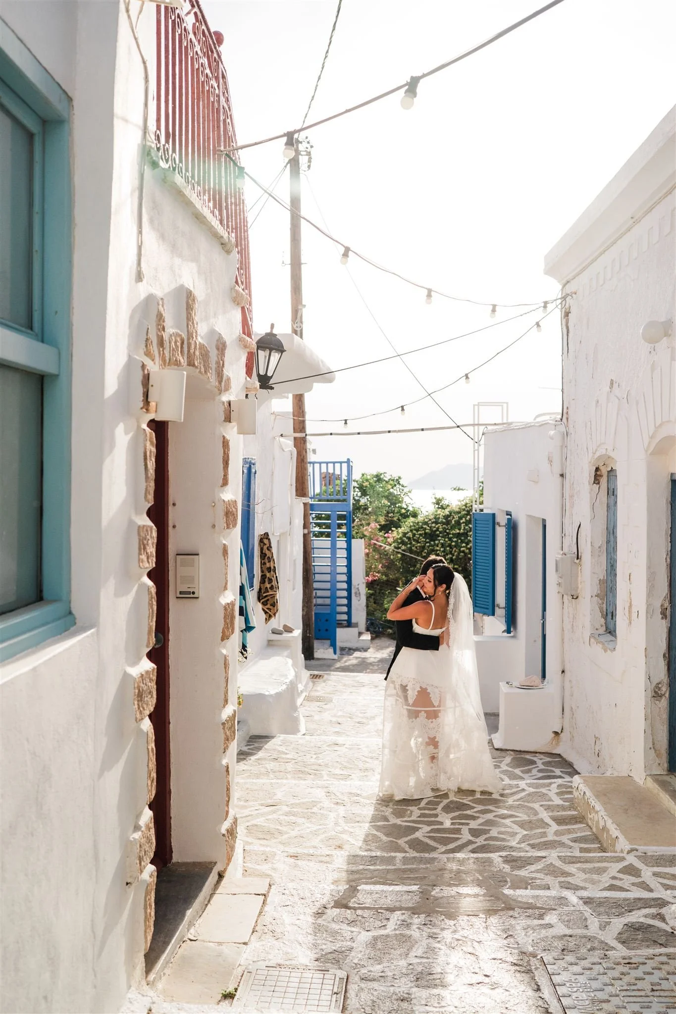 A woman in a wedding dress standing in a white stone alleyway, surrounded by white buildings with blue shutters, under string lights, with greenery in the background in Greece for their elopement