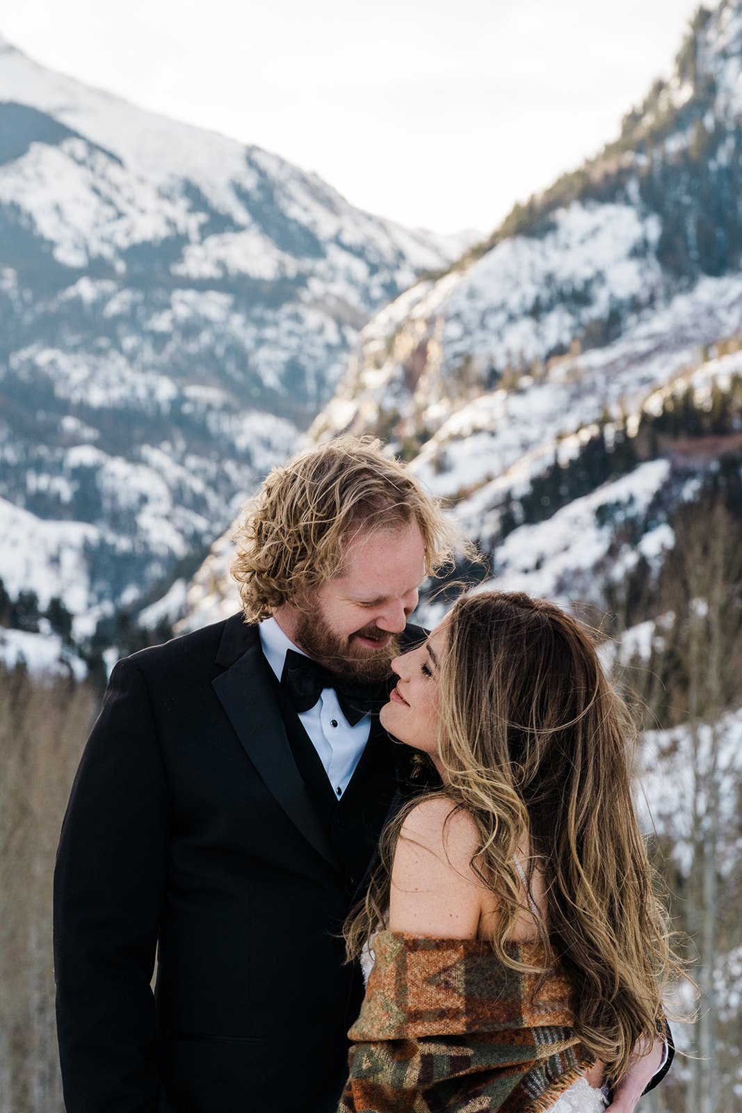 A couple dressed in wedding attire standing close together outdoors in a snowy mountain landscape, smiling and looking lovingly at each other.