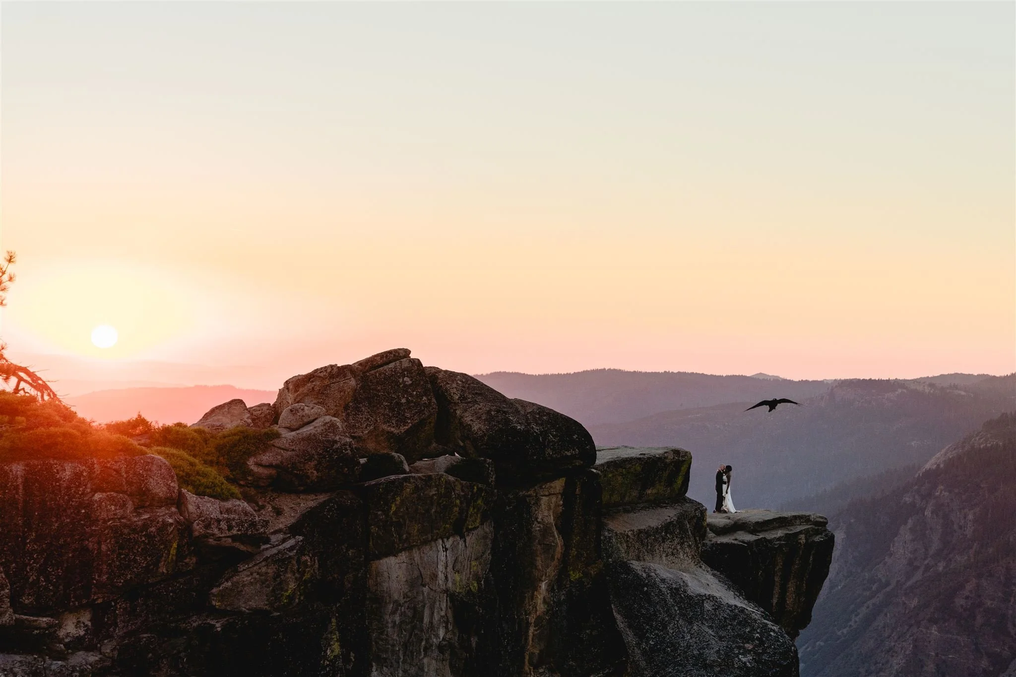 A couple dressed in wedding attire standing on a rocky cliff at sunset with mountains in the background and a bird flying nearby.