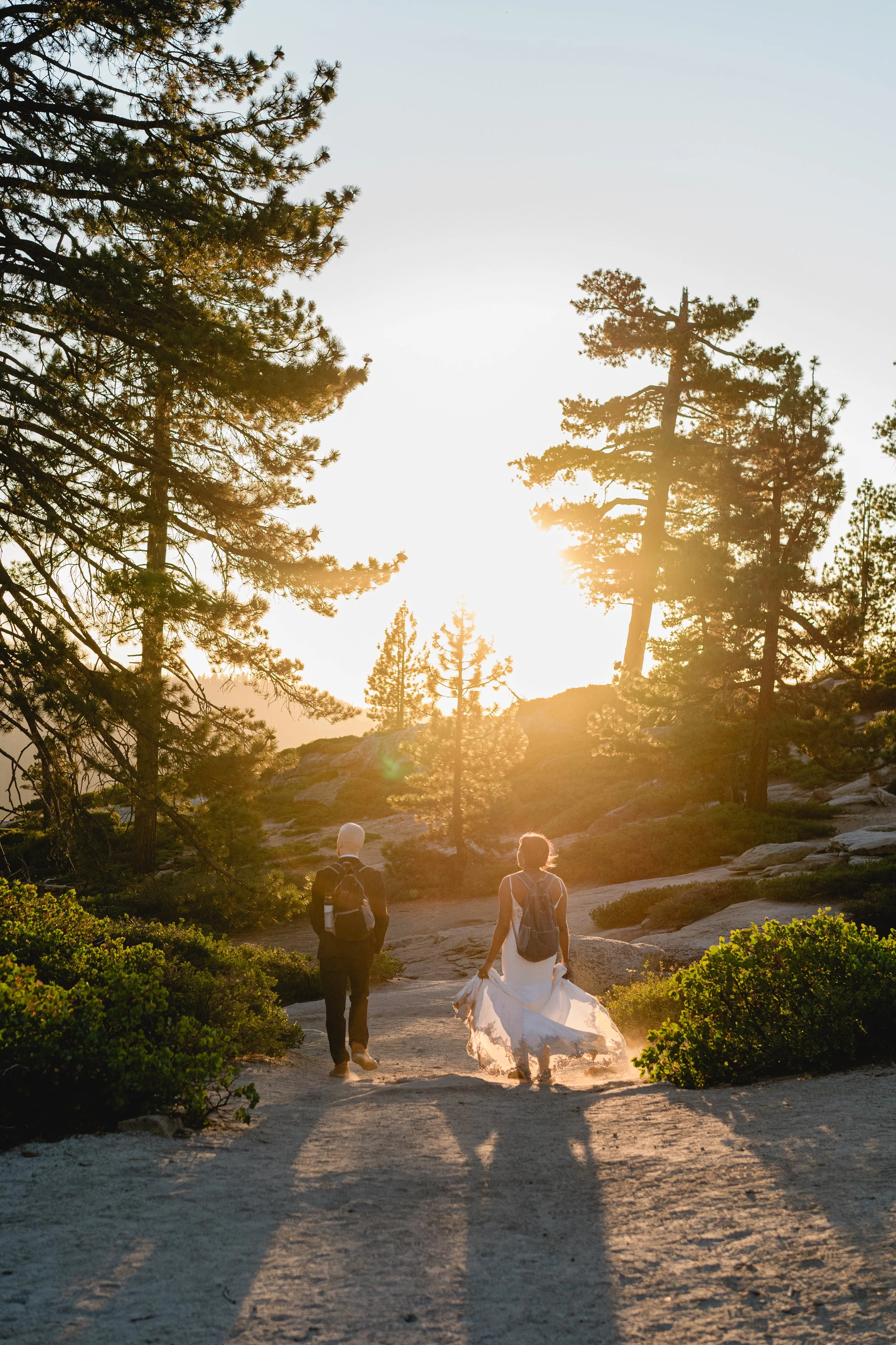 Two people walking on a dirt trail through a forested area with tall pine trees during sunset, one in a white dress and the other in dark clothing with backpacks.