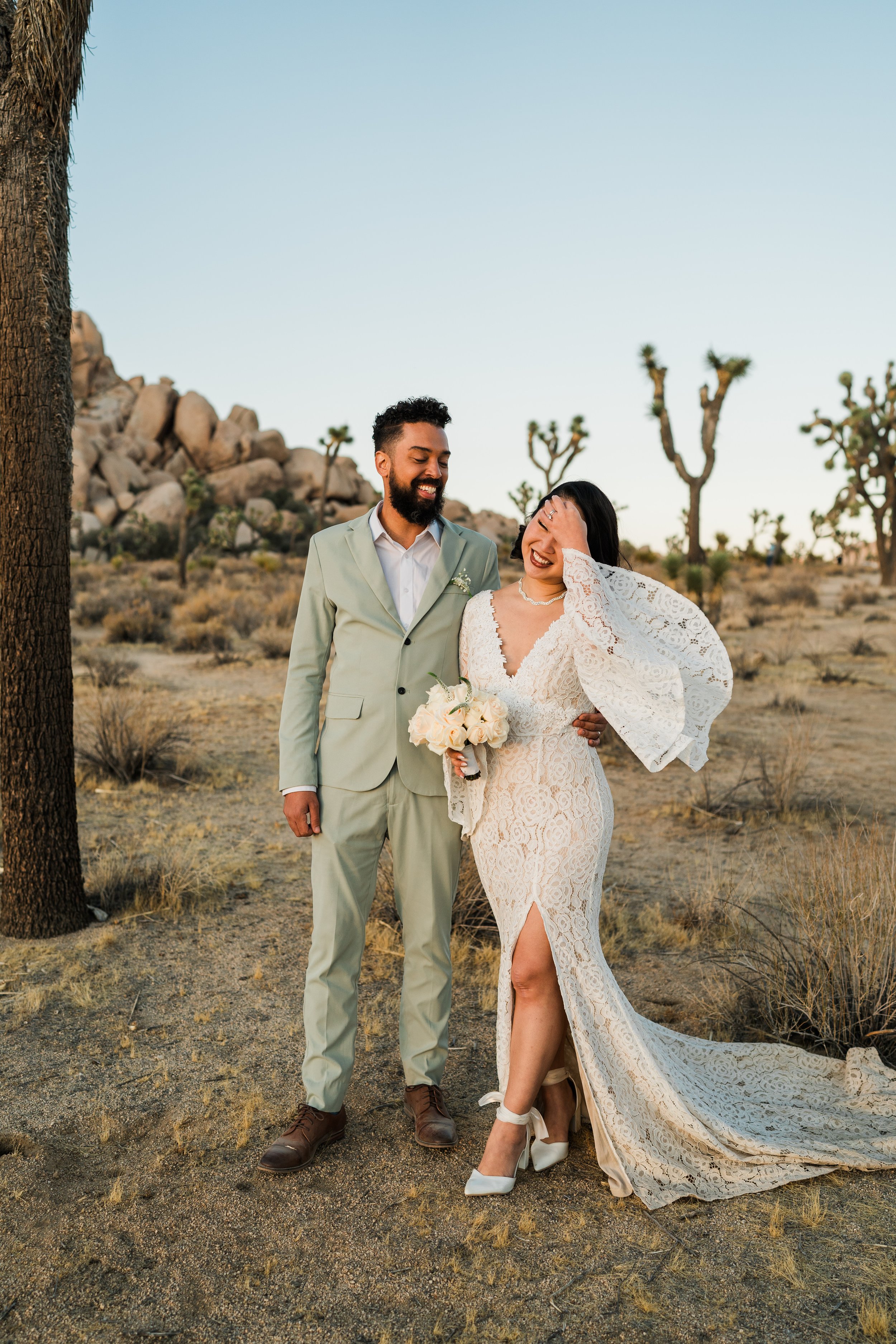 A happy couple in wedding attire standing in a desert landscape with Joshua trees and rocks, smiling and laughing together.