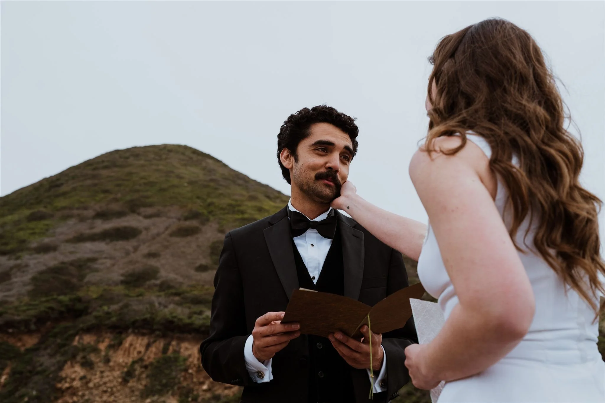 A couple having a wedding ceremony outdoors, with the groom holding a book and the bride touching his face. They are dressed in formal wedding attire, with a hill in the background.