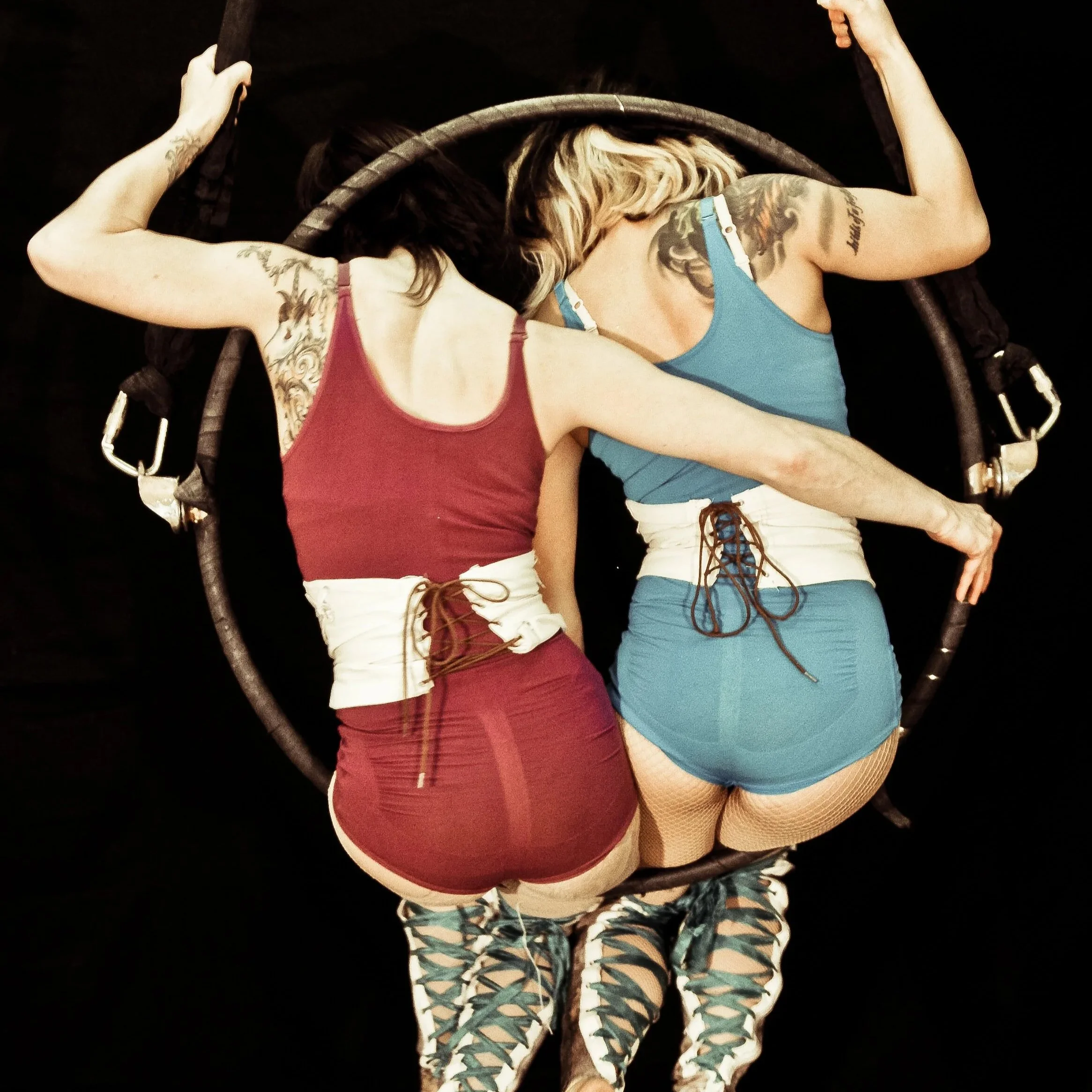 Two women sit in a hanging hoop, facing away from camera, one in red leotard, one in blue leotard