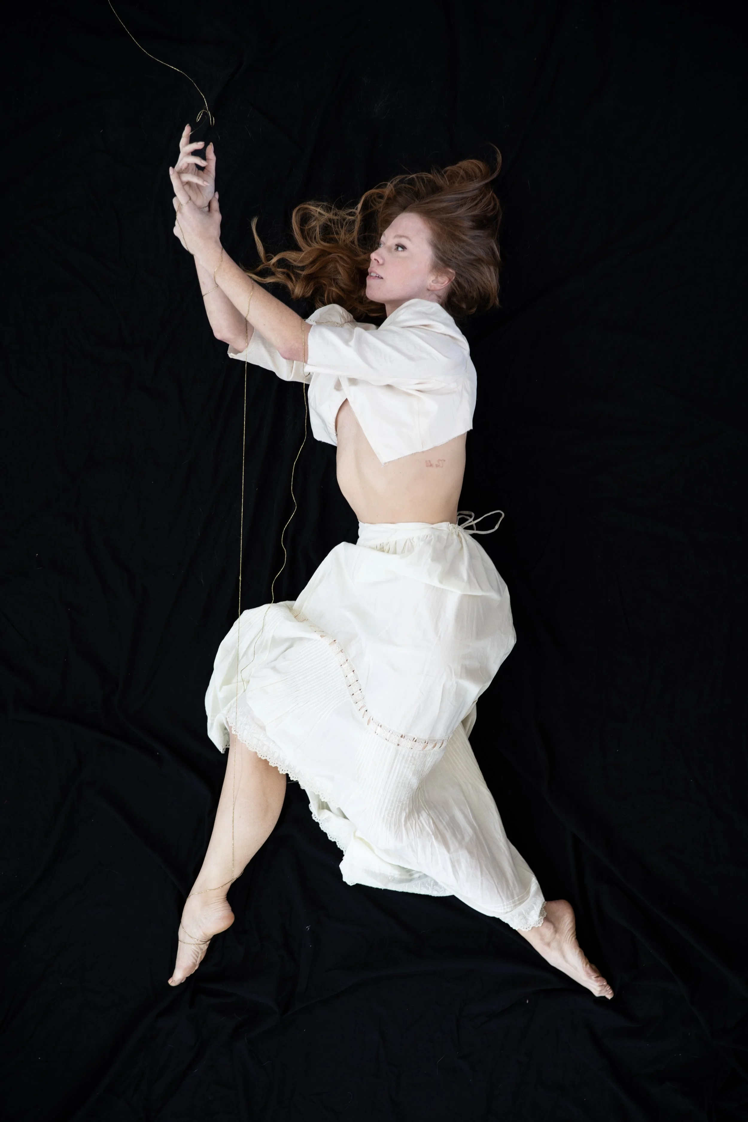 Woman in white lies against black background, hanging onto the golden thread dropping from above.