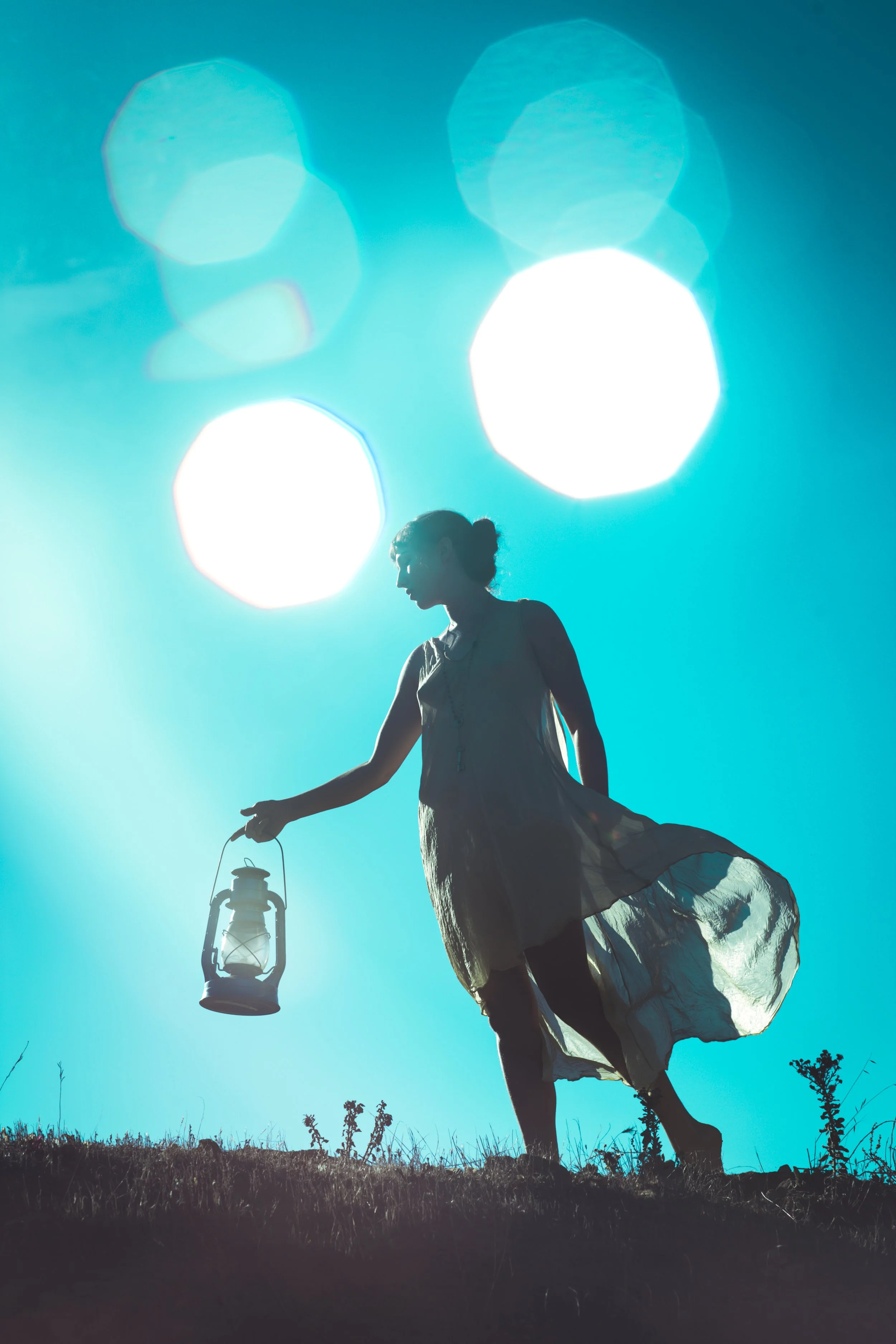 Silhouette of a woman against a bright turquoise sky, light flares near her head, she holds a lantern and appears to be in search of something