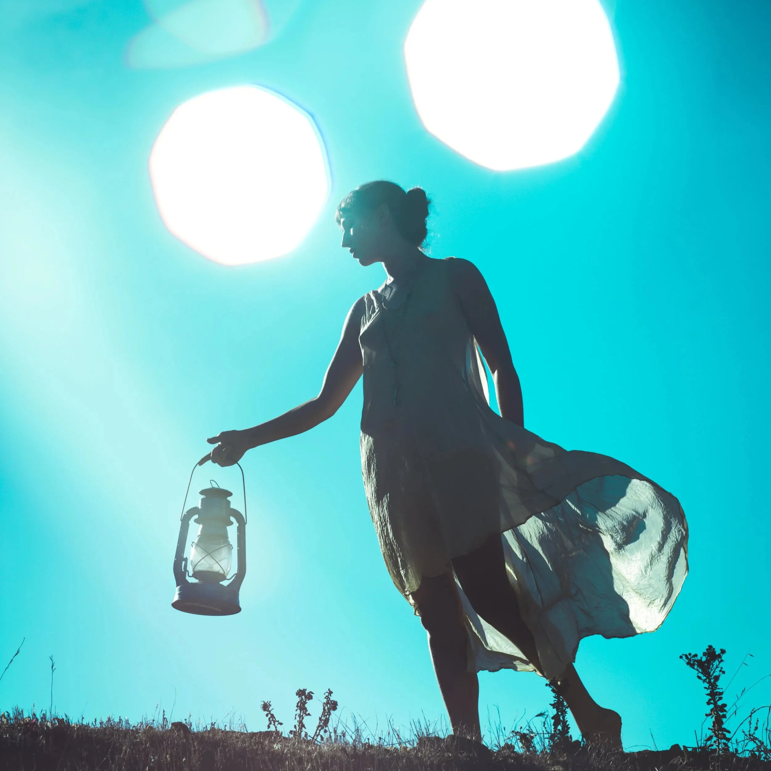 Silhouette of a woman against a bright turquoise sky, light flares near her head, she holds a lantern and appears to be in search of something