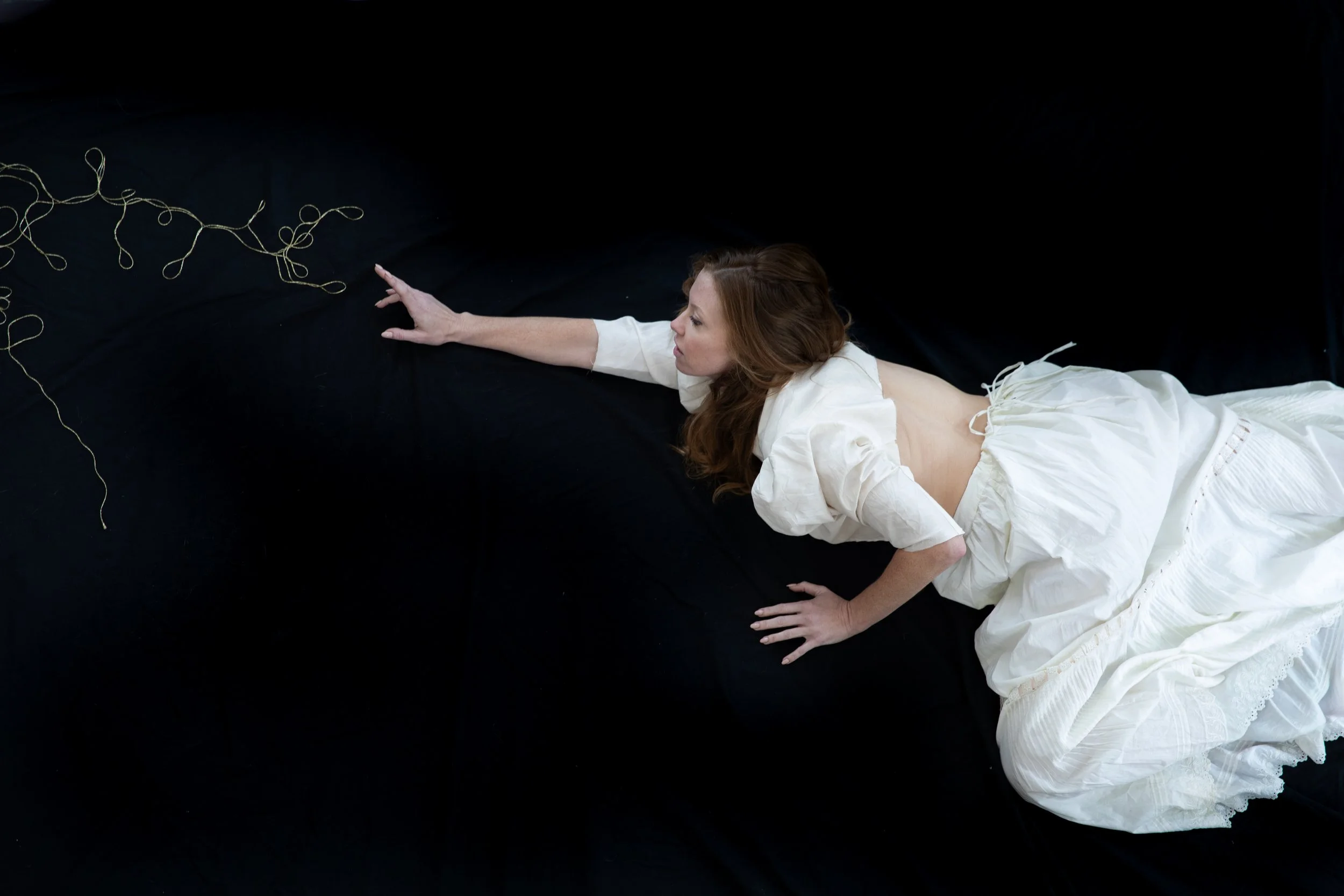 Woman in white lies across the ground, arm outstretched and reaching for the end of a golden thread.