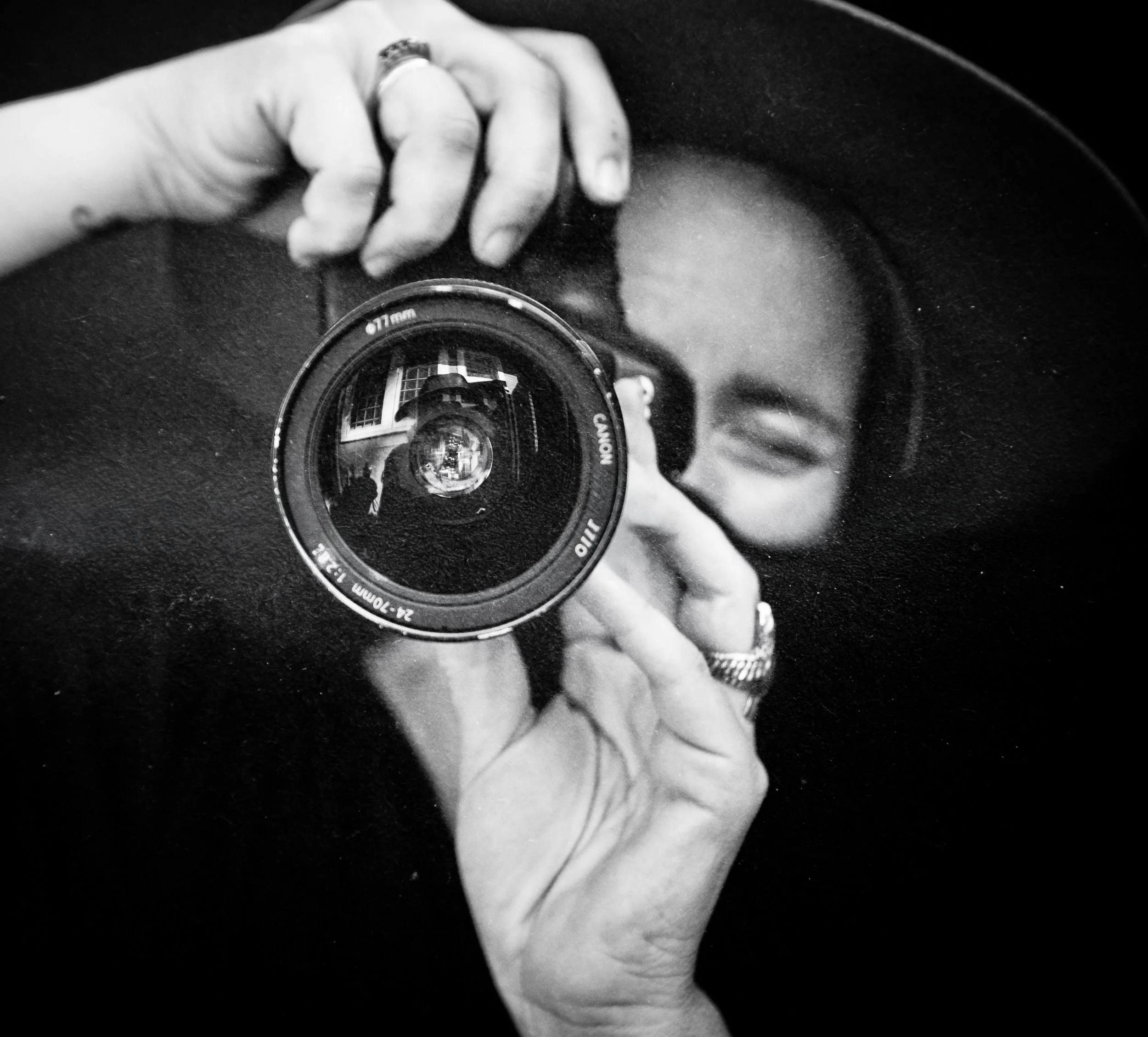 A black and white photo of a person taking a selfie in a mirror with a camera, holding the camera in front of their face, with one eye visible. The person is wearing rings on their fingers and a wristwatch.