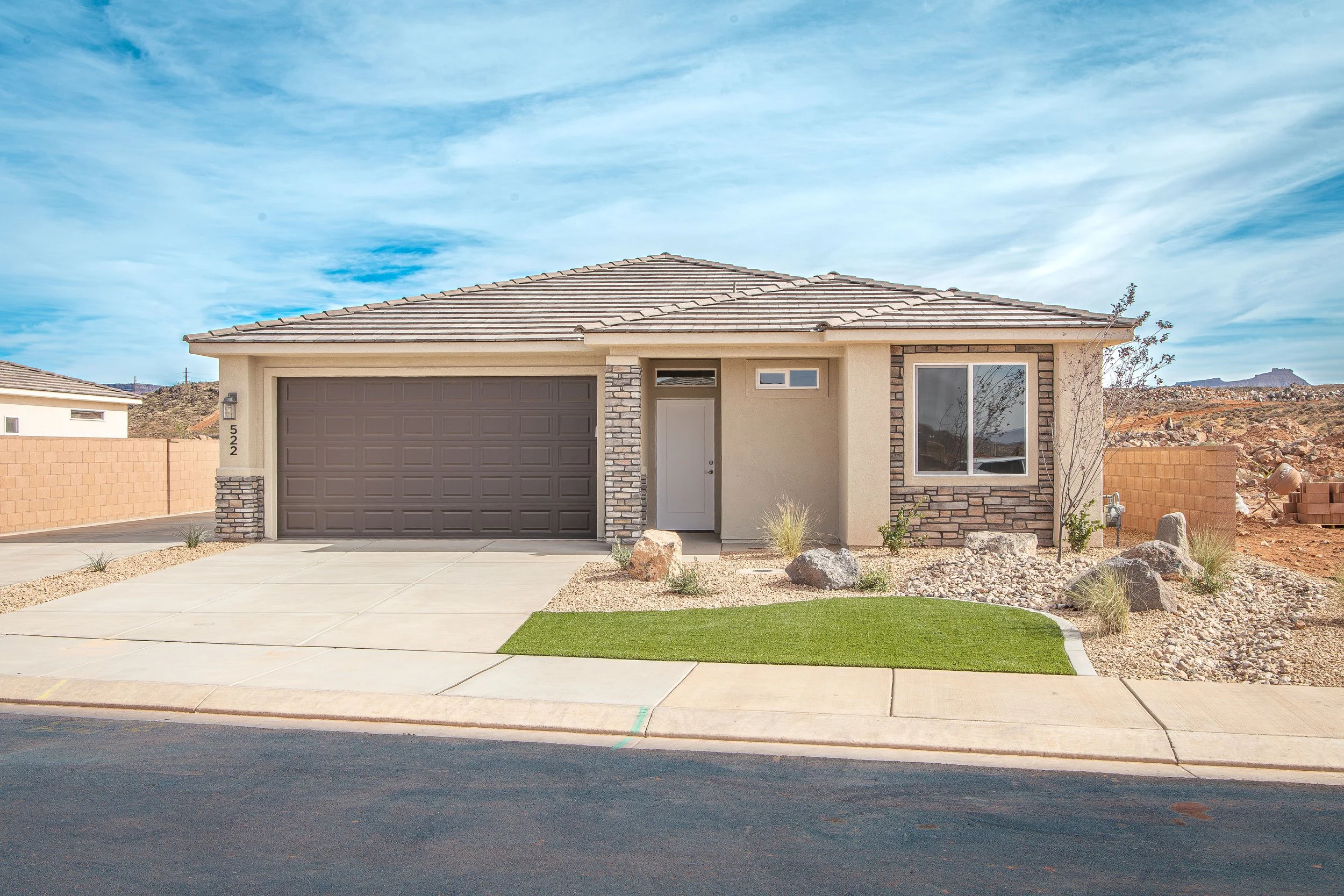 A modern single-story house with a two-car garage, a small front yard with desert landscaping, and a sidewalk in front. The house has beige walls, a tiled roof, stone accents, and a few small plants and rocks in the yard.
