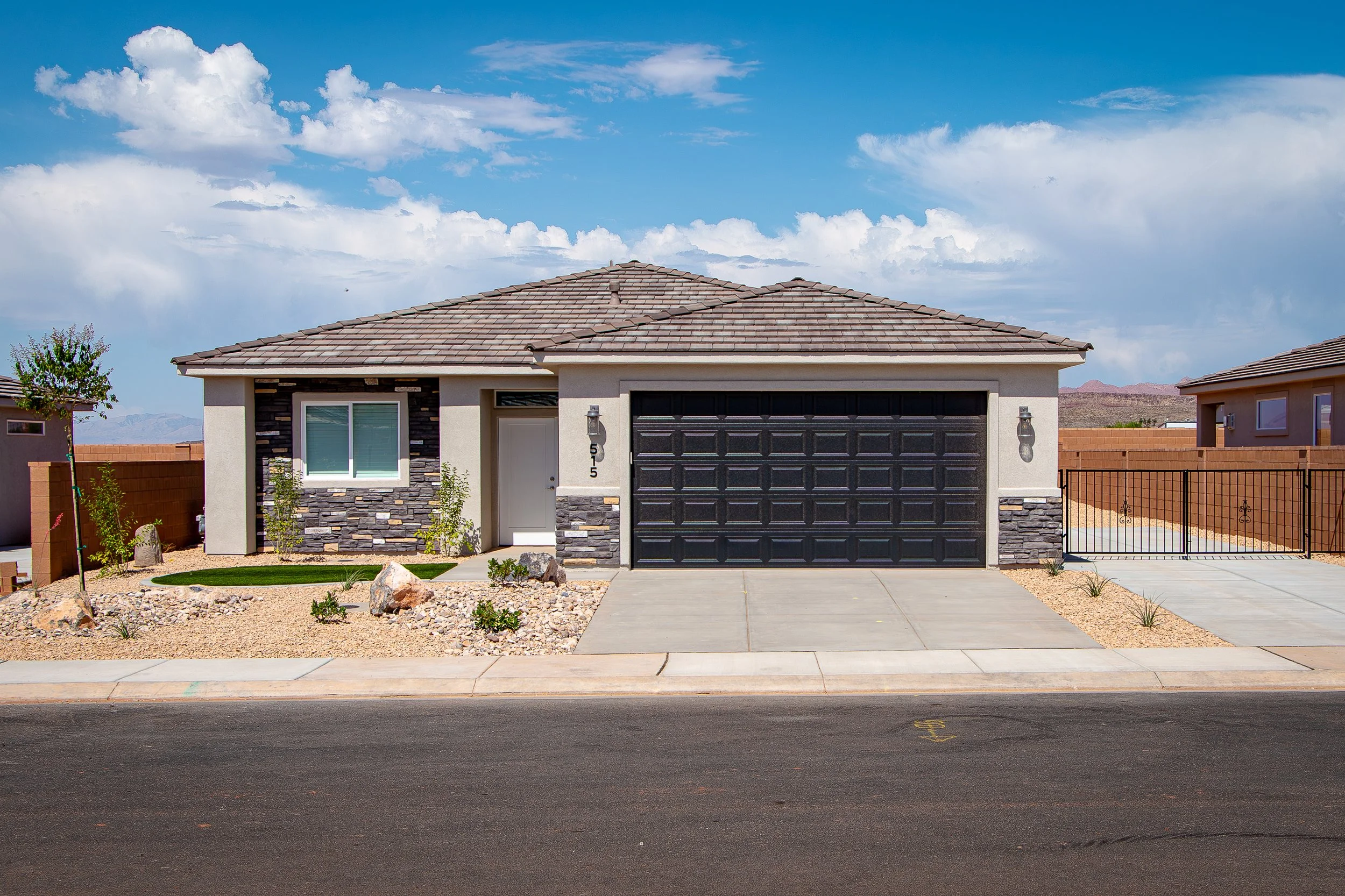 A modern single-story house with a gray and black color scheme, featuring a stone accent wall, a black garage door, and a small front yard with rocky landscaping and minimal greenery